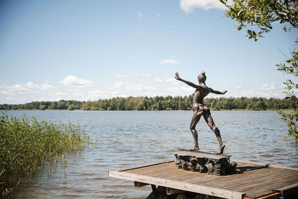 A metal sculpture of a dancing girl on a wooden dock by a lake with trees in the background and a partly cloudy sky.