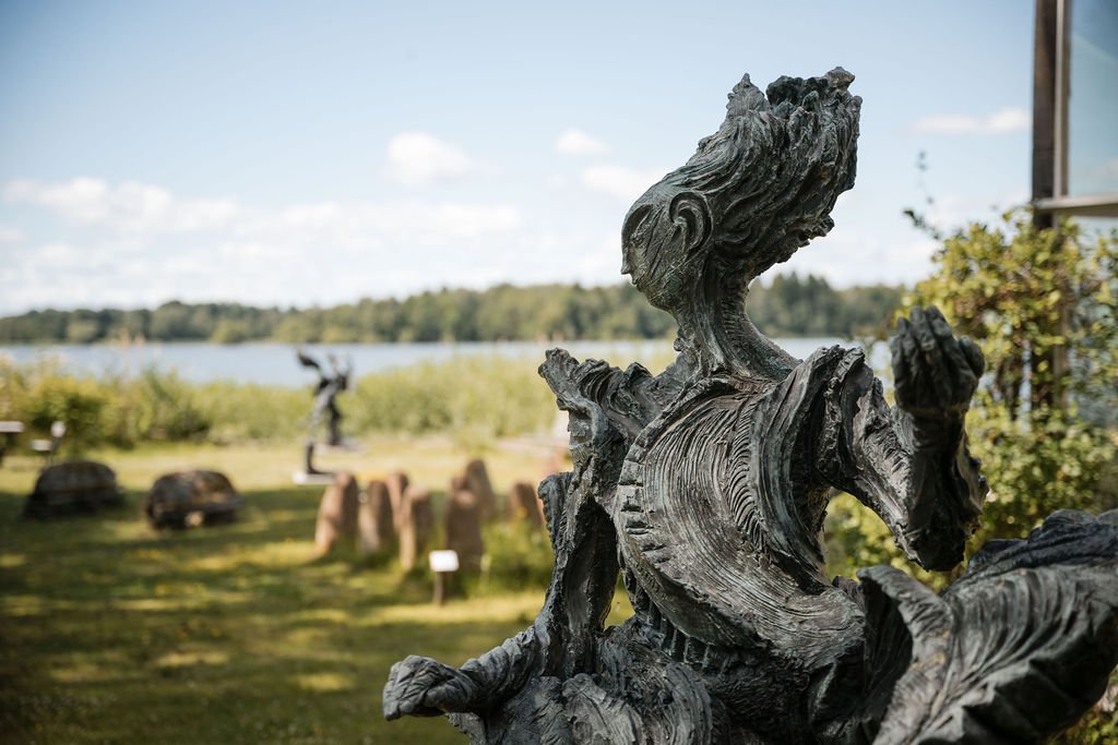 Close-up of a wooden sculpture of a woman with flowing hair, outdoors near a body of water, with other sculptures in the background.