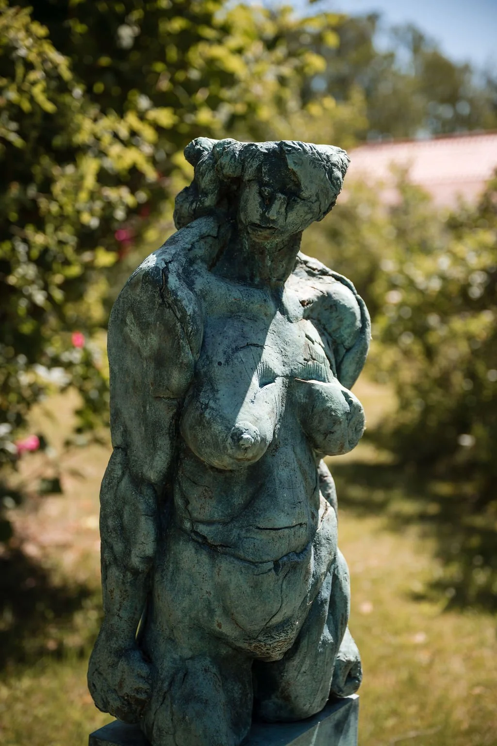 A weathered bronze statue of a nude woman with hands resting near her chest, standing outdoors with trees and a building in the background.