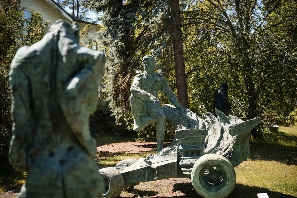 Outdoor sculpture with two human figures, one seated and one standing, and parts of a vehicle or cart, set among trees and grass.