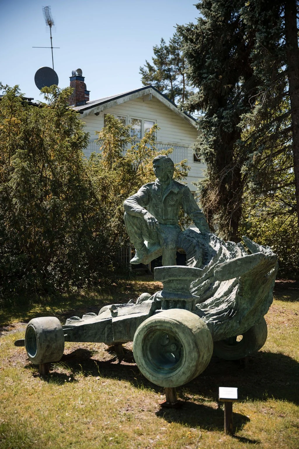 Bronze statue of a person sitting on a disassembled go-kart, outdoors with trees and a house in the background.