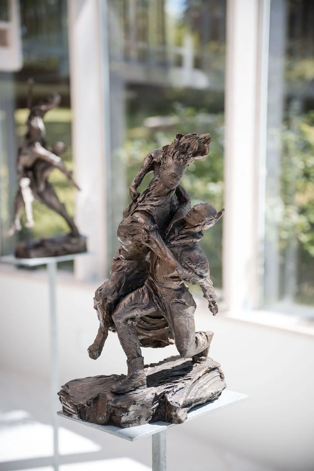 Bronze sculpture of two children playing, one child helping the other to climb, displayed on a pedestal in an art gallery.