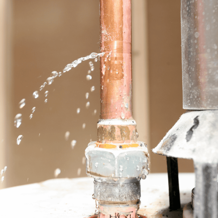 A close-up of a leaking pipe on a wet sidewalk, with orange and white traffic cones and a utility vehicle in the background