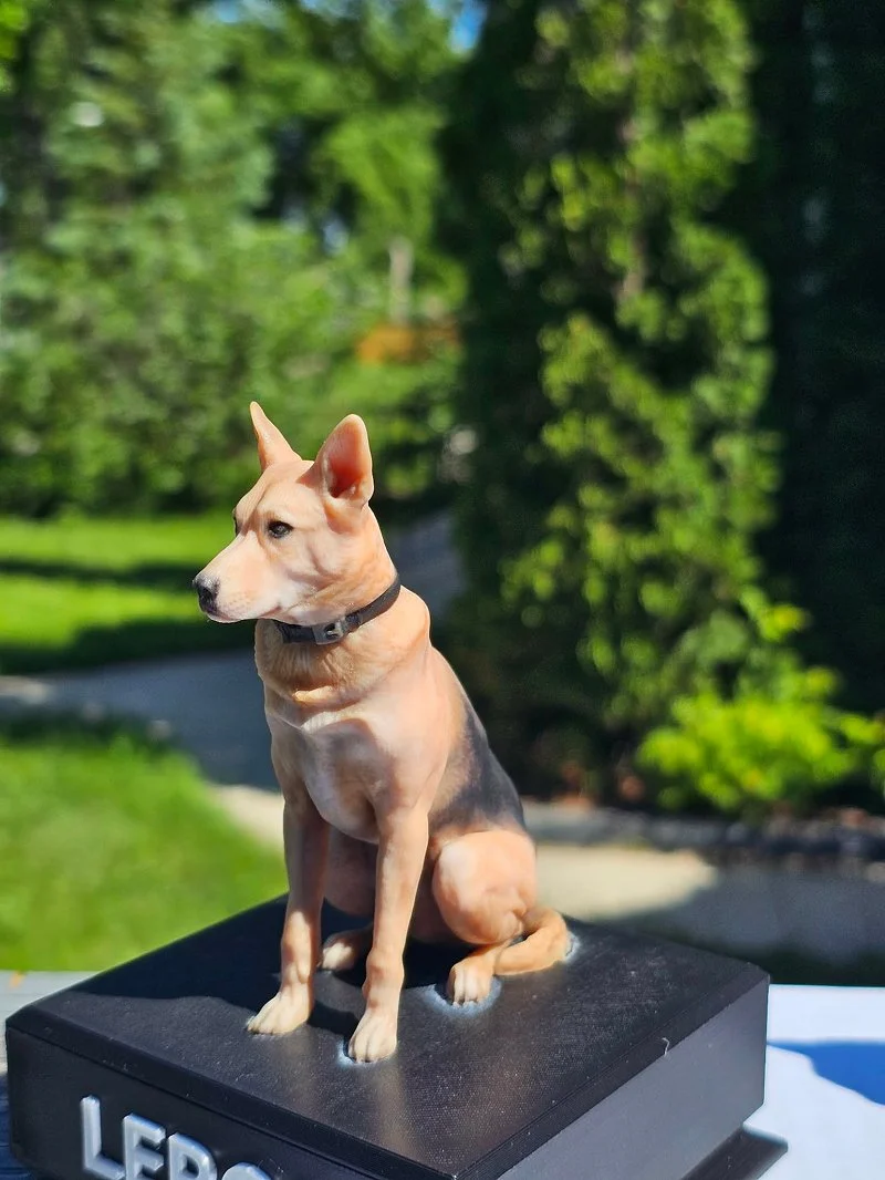 A realistic sculpture of a young dog, possibly a pitbull or similar breed, sitting on a black rectangular platform outdoors with green trees and a pathway in the background.