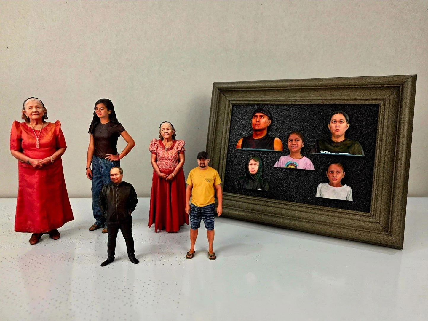 A group of seven miniature figures resembling people, arranged on a white surface with a plain white background. To the right, a large framed picture displays six miniature faces. The figures include women, men, and children, dressed in casual and traditional clothing.