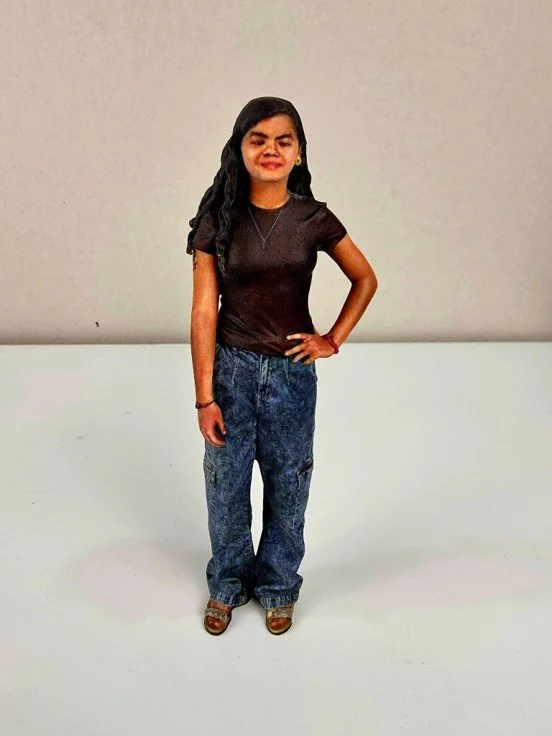 A woman with dark wavy hair standing against a plain wall, wearing a black top, blue jeans, and brown heels, with her right hand on her hip.