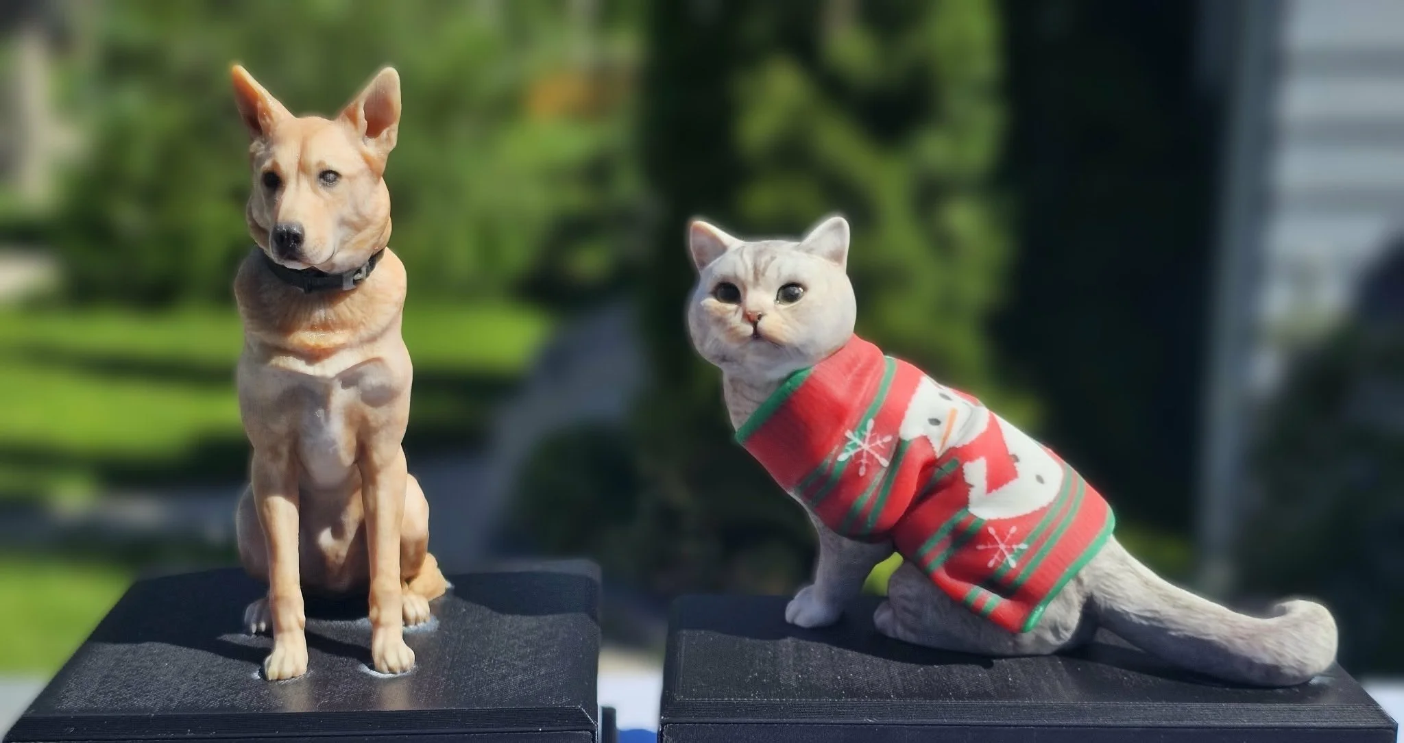 A dog with a tan coat and pointed ears sitting on a display stand, and a cat with a light-colored coat wearing a festive red and green sweater with snowflake and reindeer patterns, sitting on a similar display stand, both posed outdoors with blurred green trees in the background.