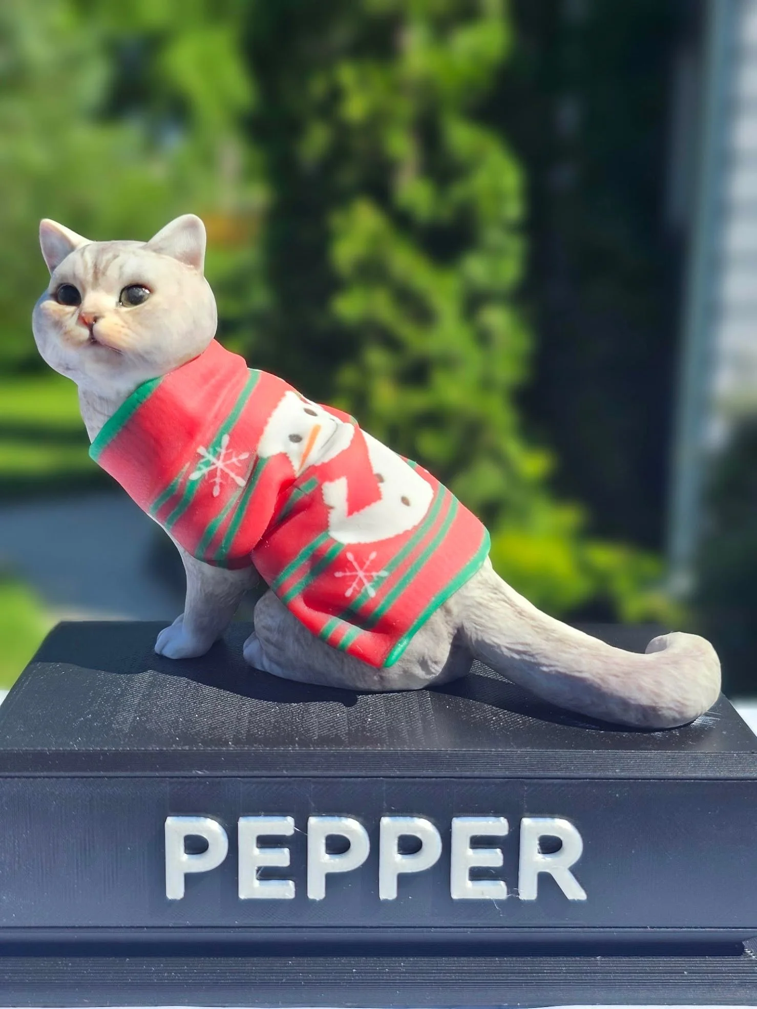 A cat figurine dressed in a red holiday sweater with a snowman design, placed on a black platform with the words 'PEPPER' in white letters.