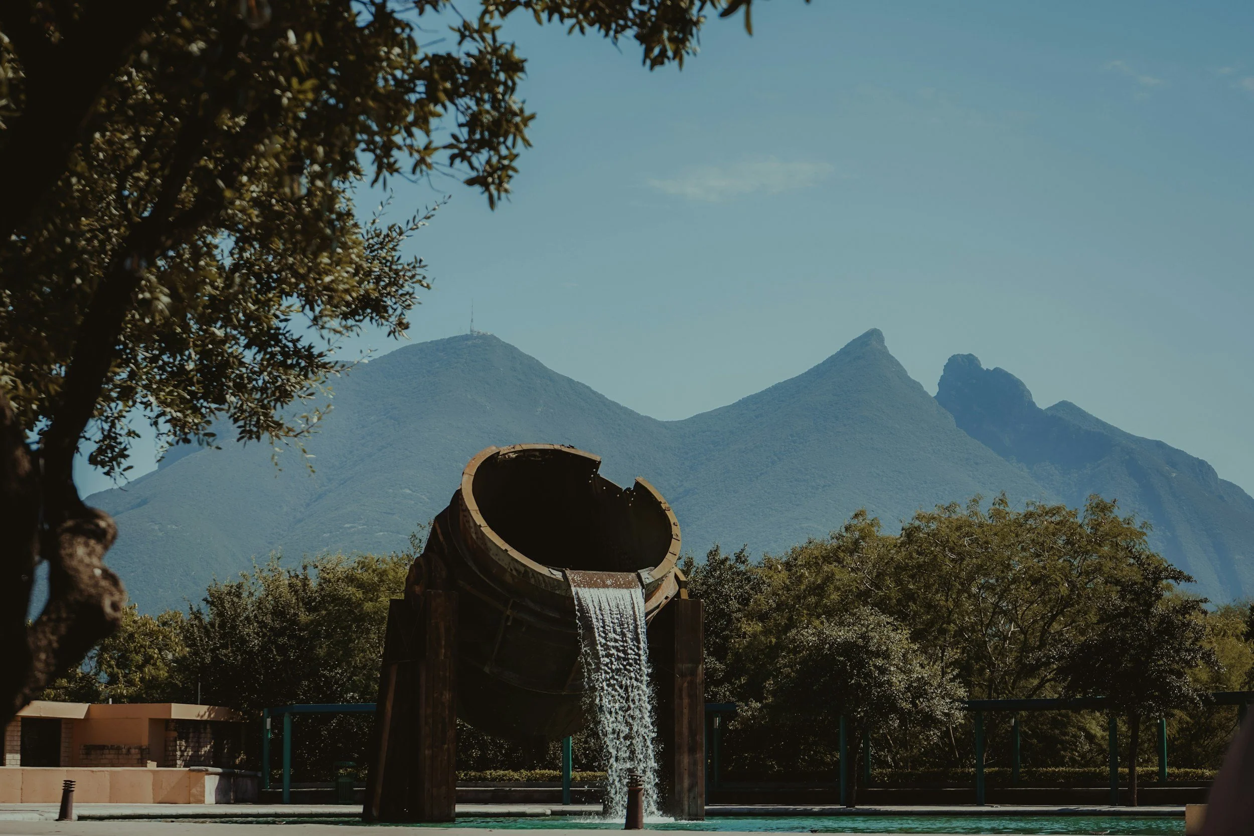 Fuente ornamental con agua fluyendo, árboles verdes y montañas en el fondo, cielo despejado.