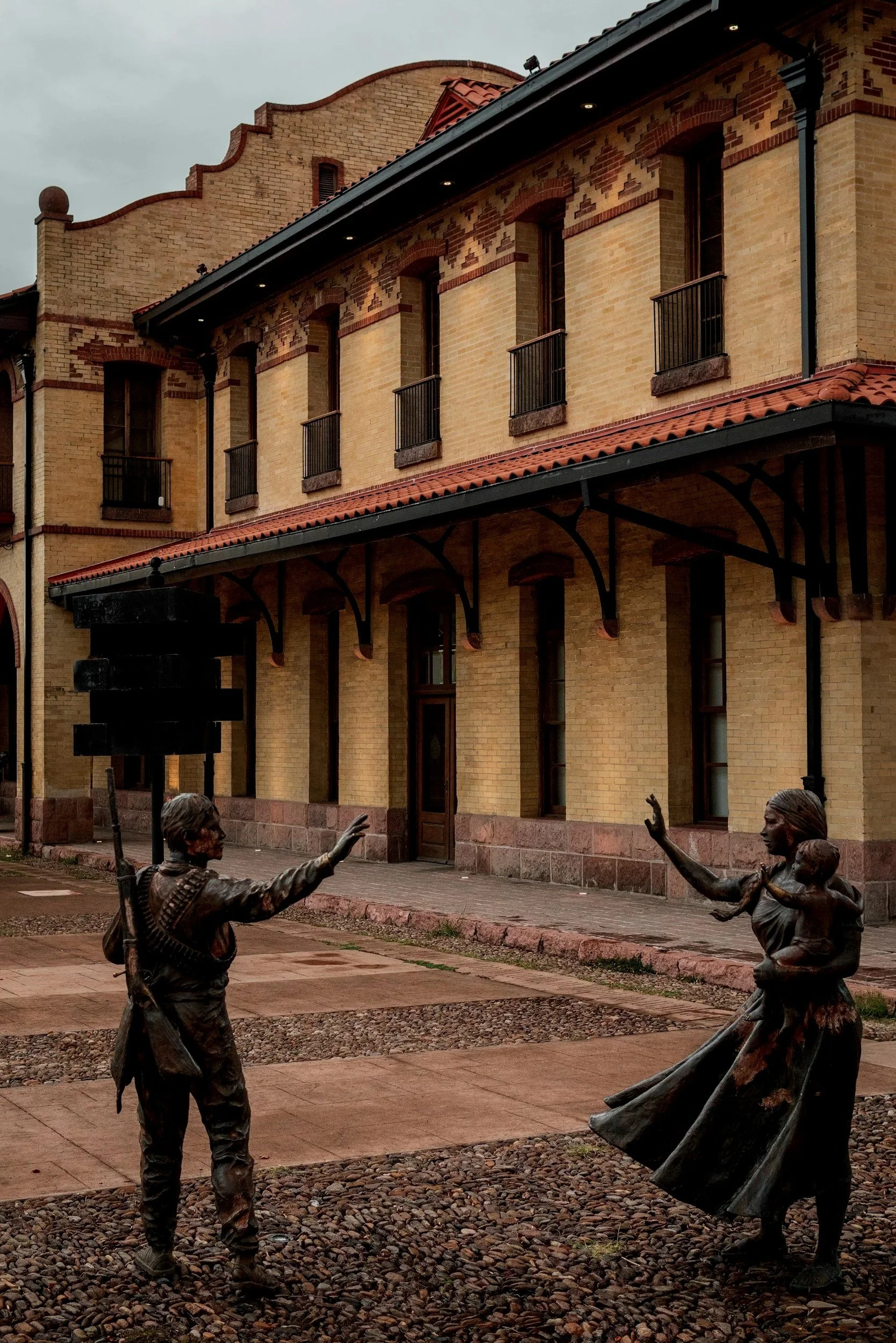 Esculturas de un hombre con rifle y una mujer con un niño en un área exterior frente a un edificio de ladrillos.