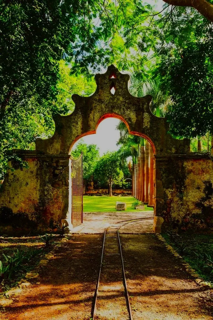 Puerta antigua en arco en medio de un jardín con árboles y rieles de tren en el suelo