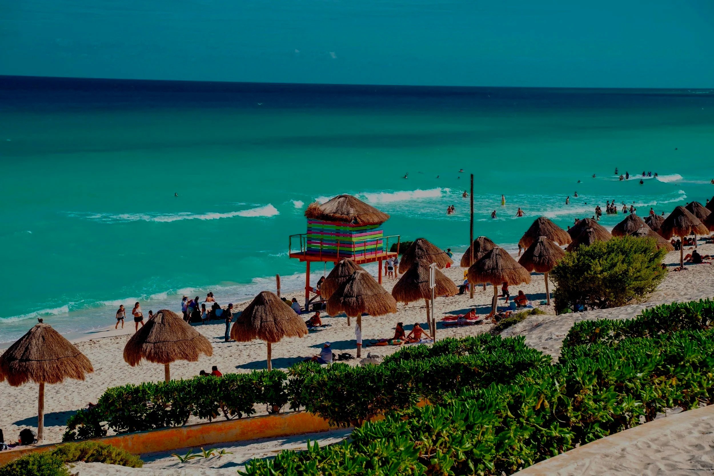 Playa con sombrillas de paja, personas en la arena y en el mar, y un puesto de vigilancia con techo colorido, en un día soleado.