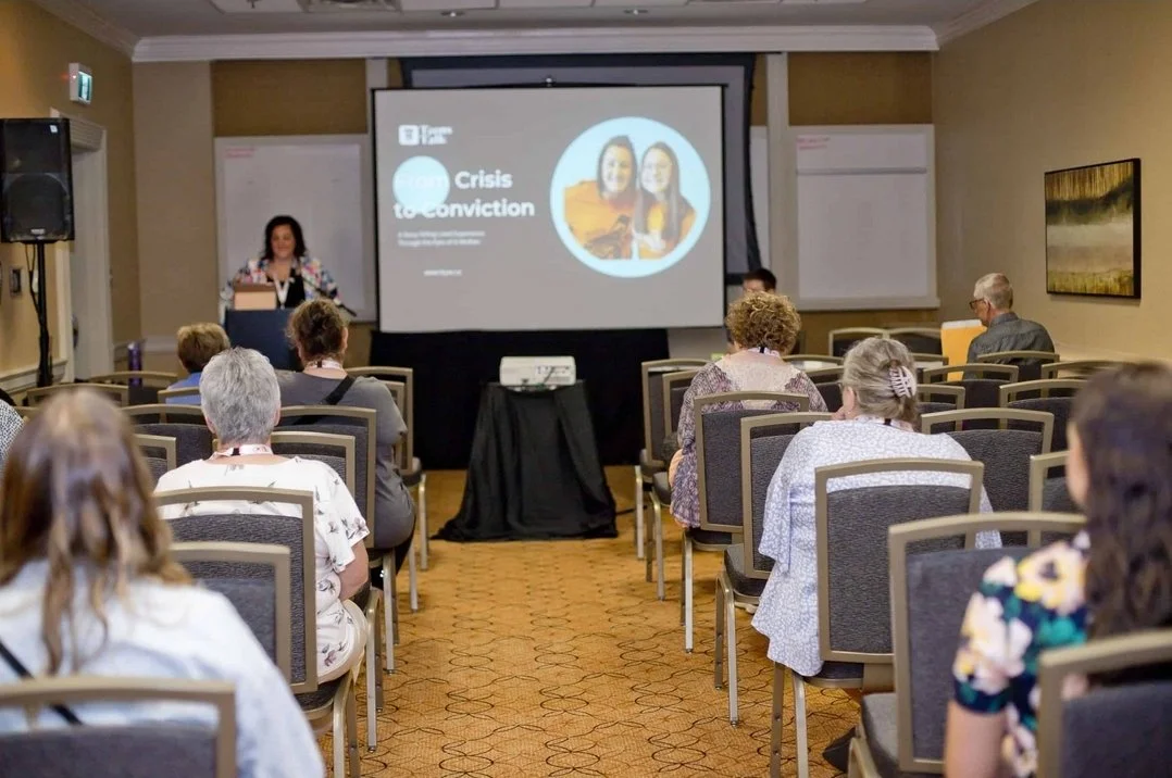 A woman presenting at a conference in a hotel meeting room with an audience seated facing the speaker and a projector screen displaying a slide titled "Crisis to conviction."