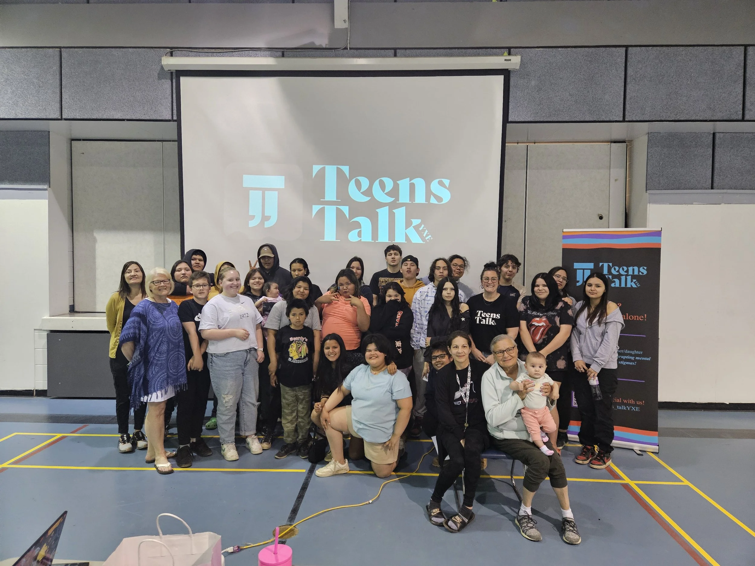 Group of people standing in front of a large screen and a banner at a Teen Talks event, inside a gymnasium.