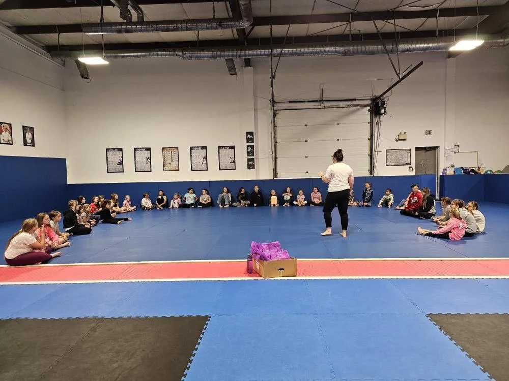 A group of children sitting in a large circle on a blue padded floor inside a martial arts or gym training facility, listening to an instructor in white standing in the center.