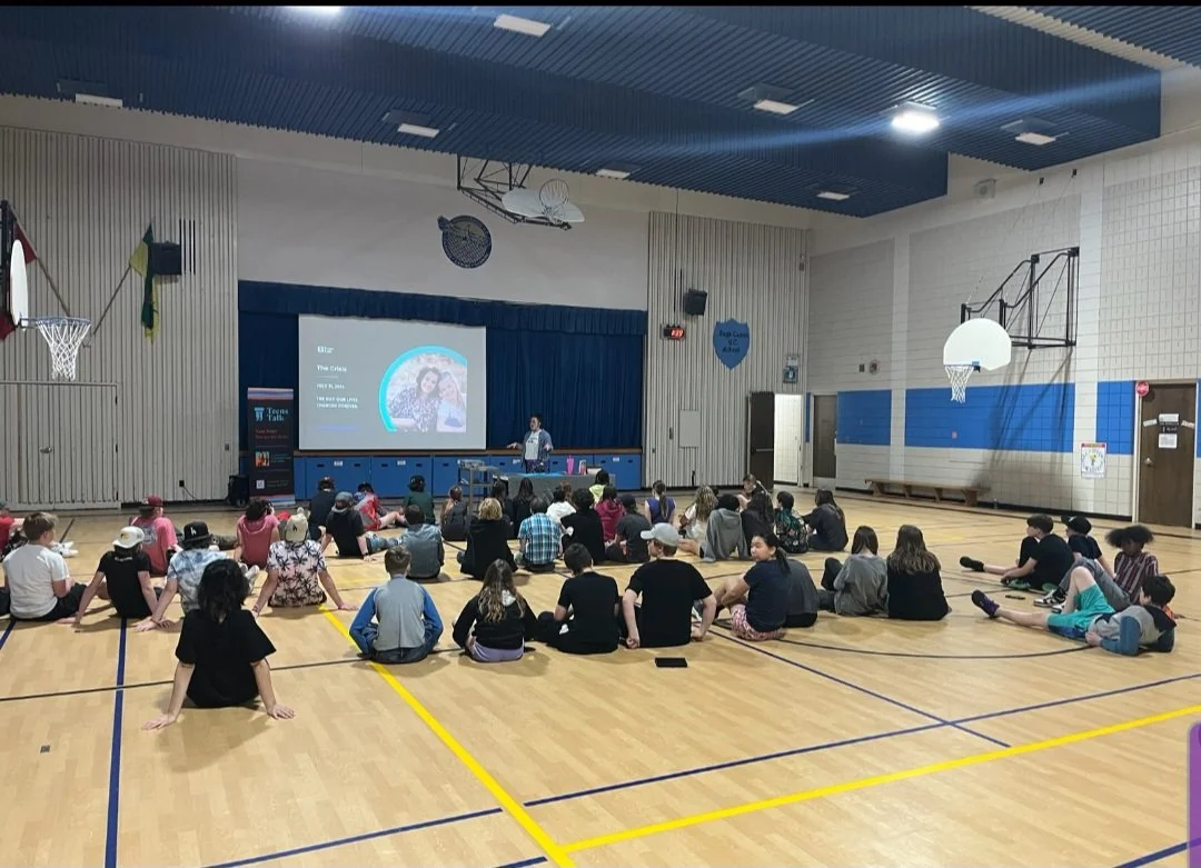 Students sitting on the gymnasium floor watching a presentation on a large screen, with a speaker standing at a podium in front.