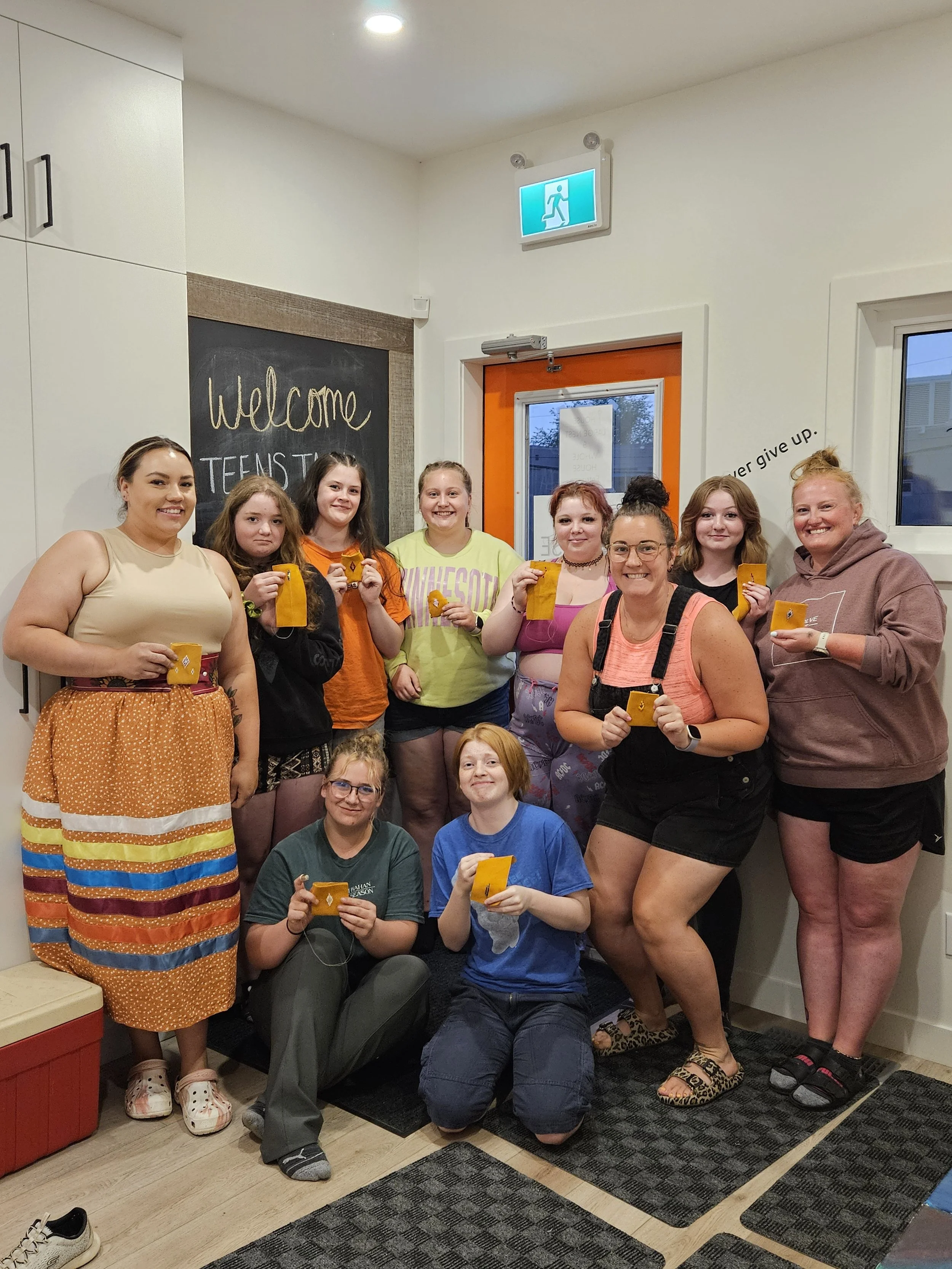 A group of ten young women and girls posing together indoors, holding orange-colored items, in a room with a chalkboard that reads 'Welcome Teen' and an emergency exit sign above a door.