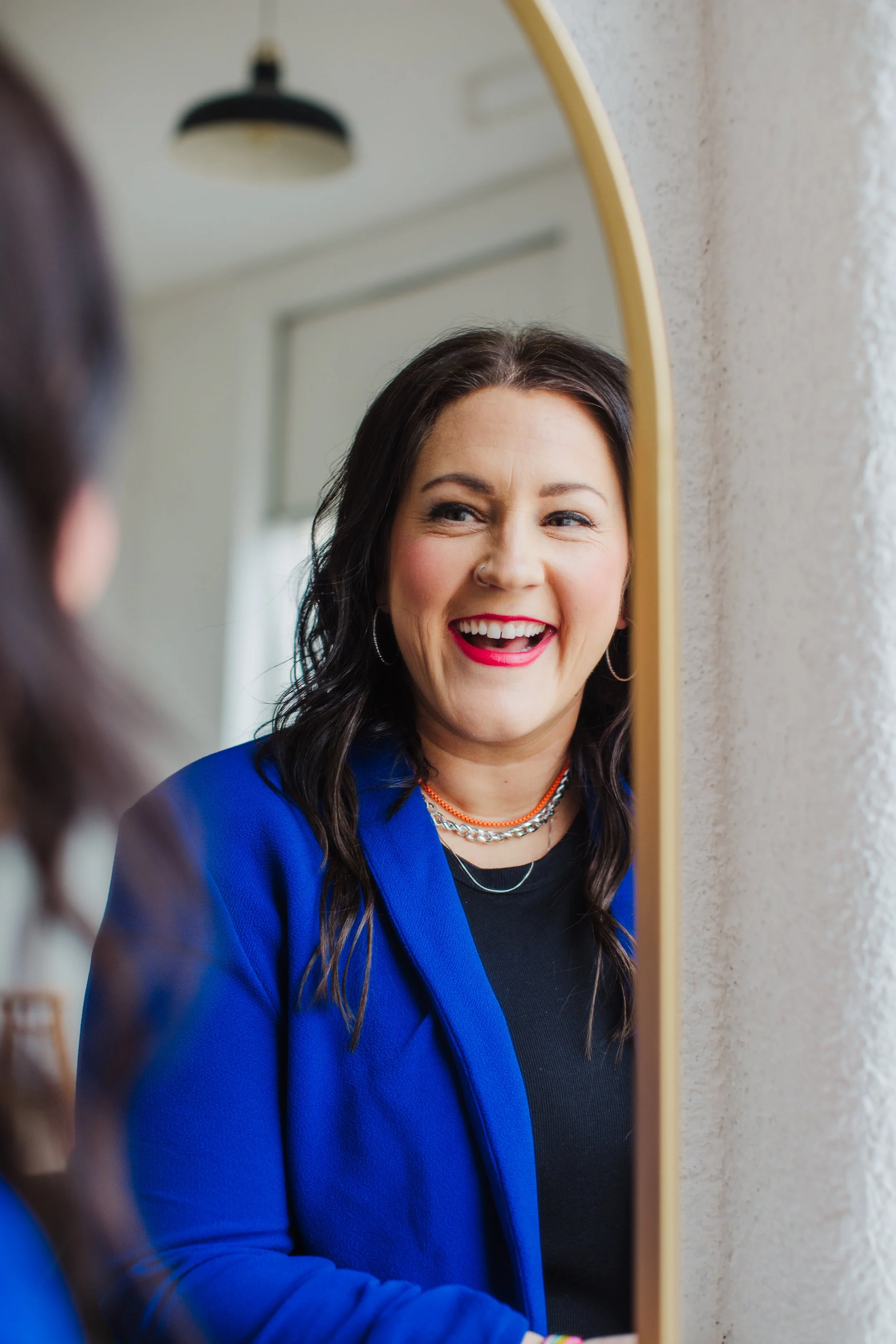 A woman with dark hair, wearing a blue blazer and gold jewelry, looking at her reflection in a mirror with a big smile.
