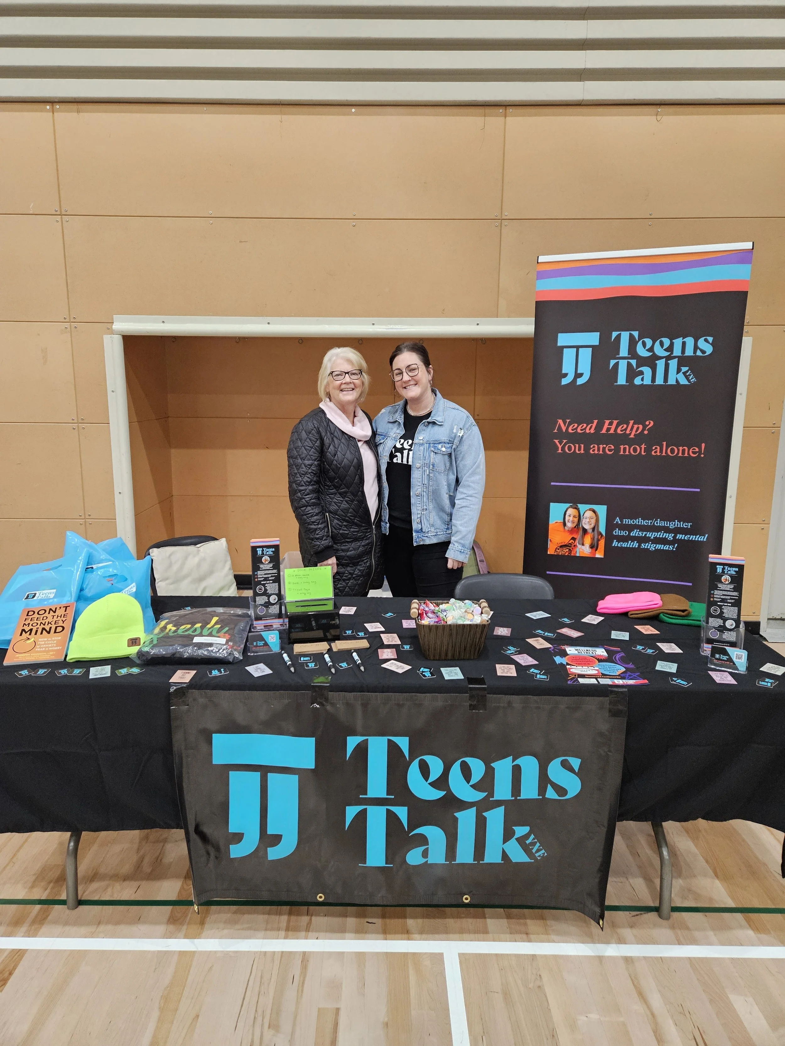 Two women standing behind a table at a Teen Talk informational booth, with a banner and various pamphlets and items on the table.