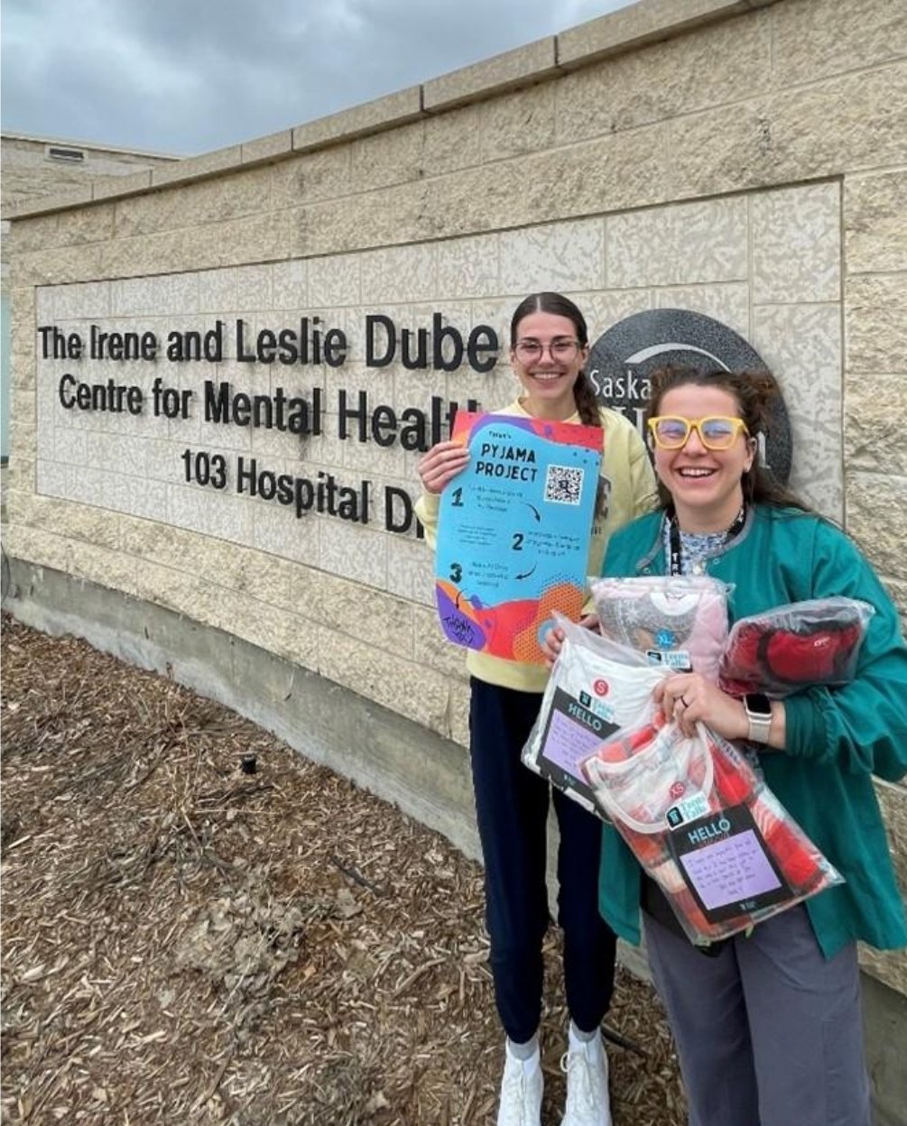 Two women standing outside the Irane and Leslie Dube Centre for Mental Health at a hospital, holding colorful posters and packaged clothing items, smiling at the camera.