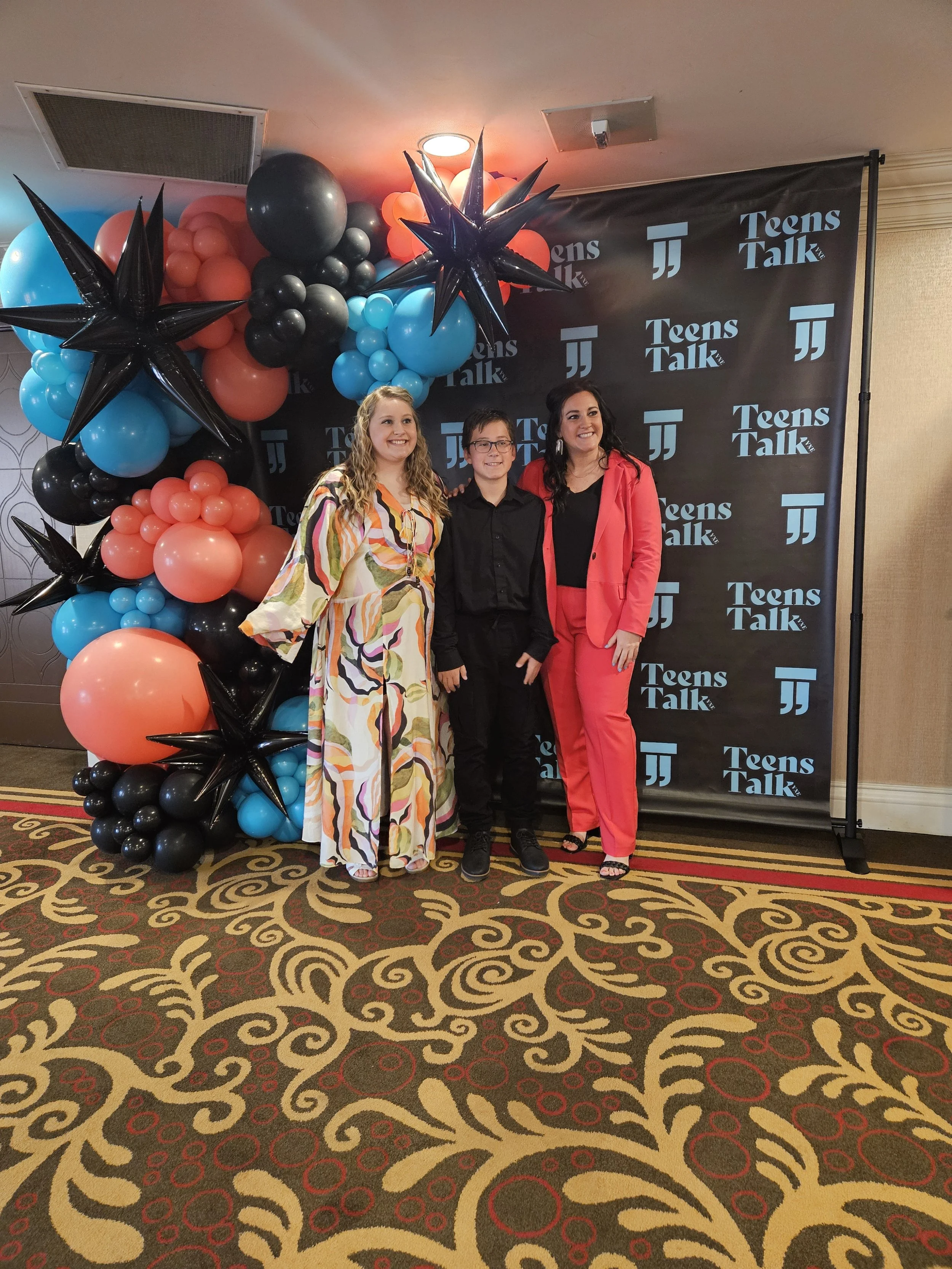 Three people posing in front of a black backdrop with blue 'Teens Talk' text and logo, surrounded by balloon decorations with star-shaped black balloons and clusters of blue, black, and coral balloons.