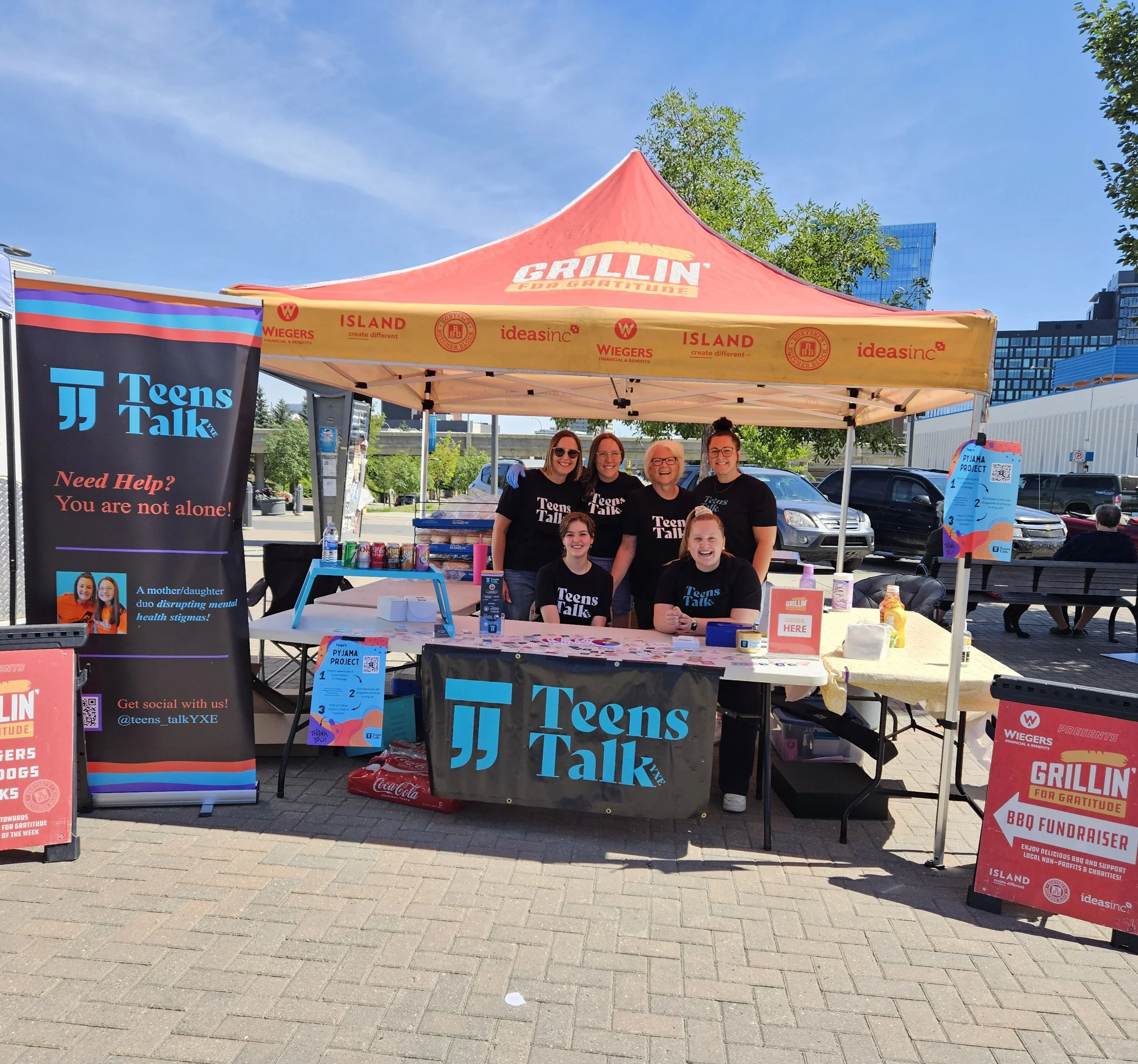 Group of five women and two girls under a yellow and red canopy with 'Grillin for Gratitude' branding, at a beams and table with 'Teens Talk' banners, participating in a BBQ fundraiser event on a sunny day in an urban park.
