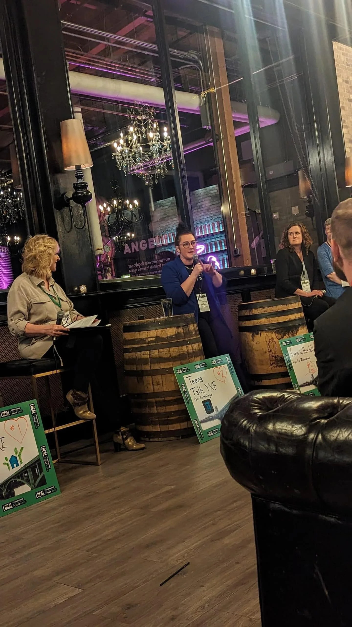A panel discussion taking place indoors with four women seated against a large window. One woman in a blue blazer is speaking into a microphone. The setting has chandeliers, exposed brick, and dim lighting. There are signs on the floor indicating topics like 'Teens Talk XYE' and 'Farm to Table'.