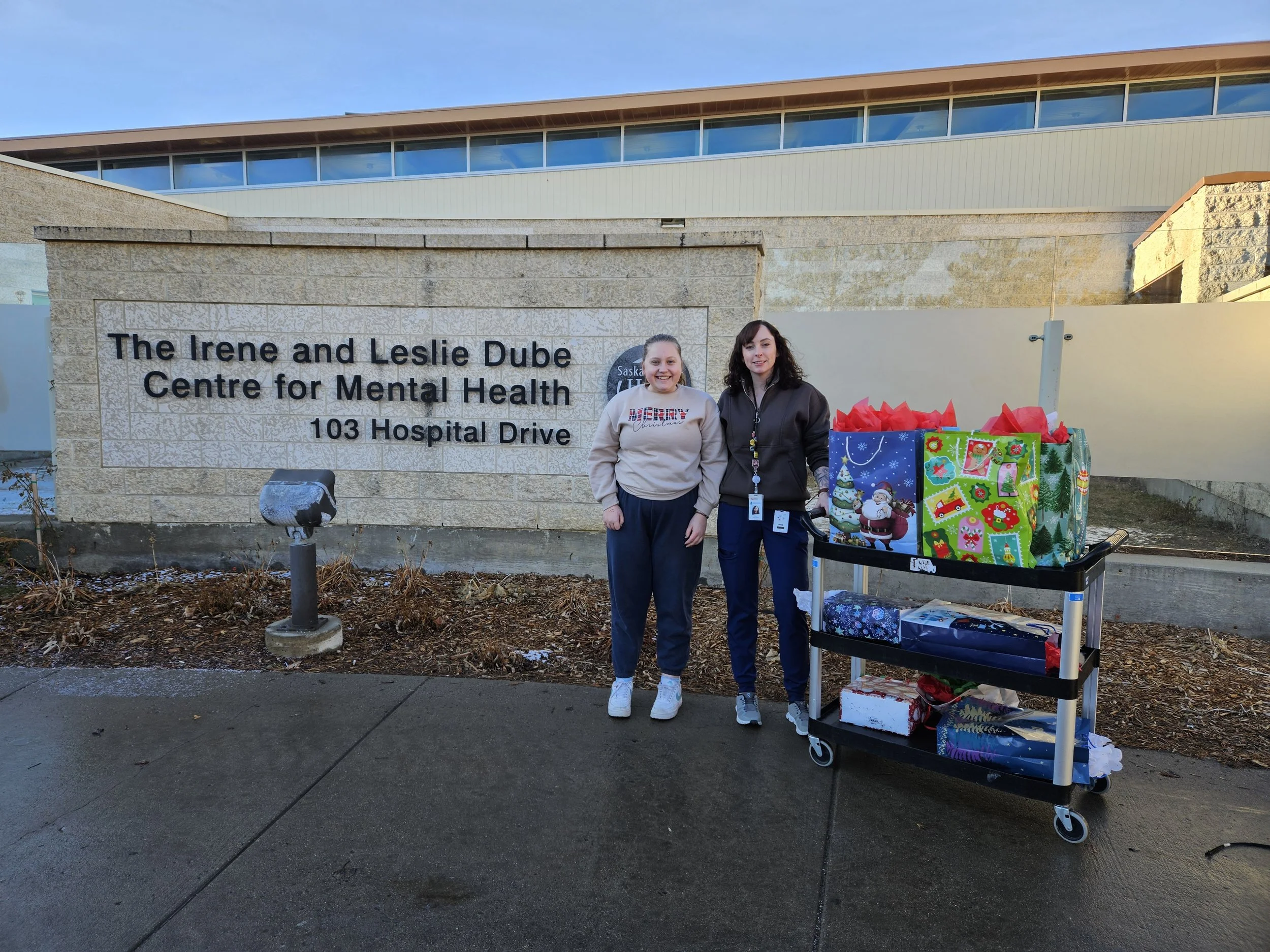 Two women standing outside The Irene and Leslie Dube Centre for Mental Health, with a cart of wrapped presents for Christmas.