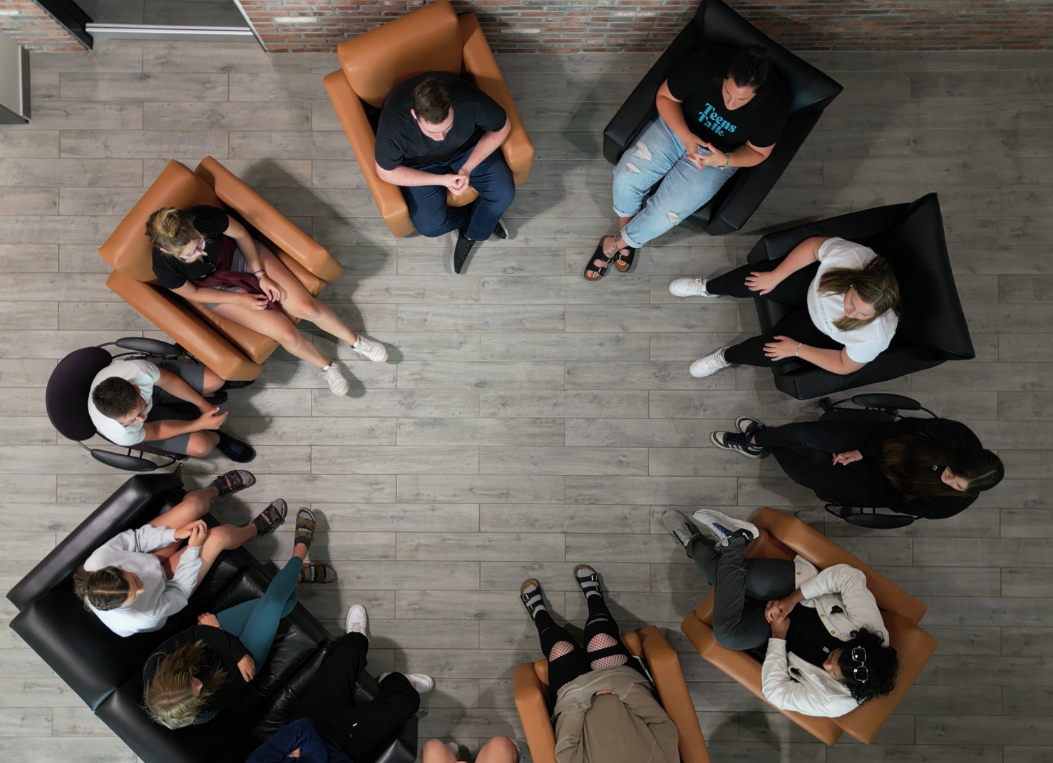A group of people sitting in a circle in a room with wooden flooring, engaged in a discussion or meeting.