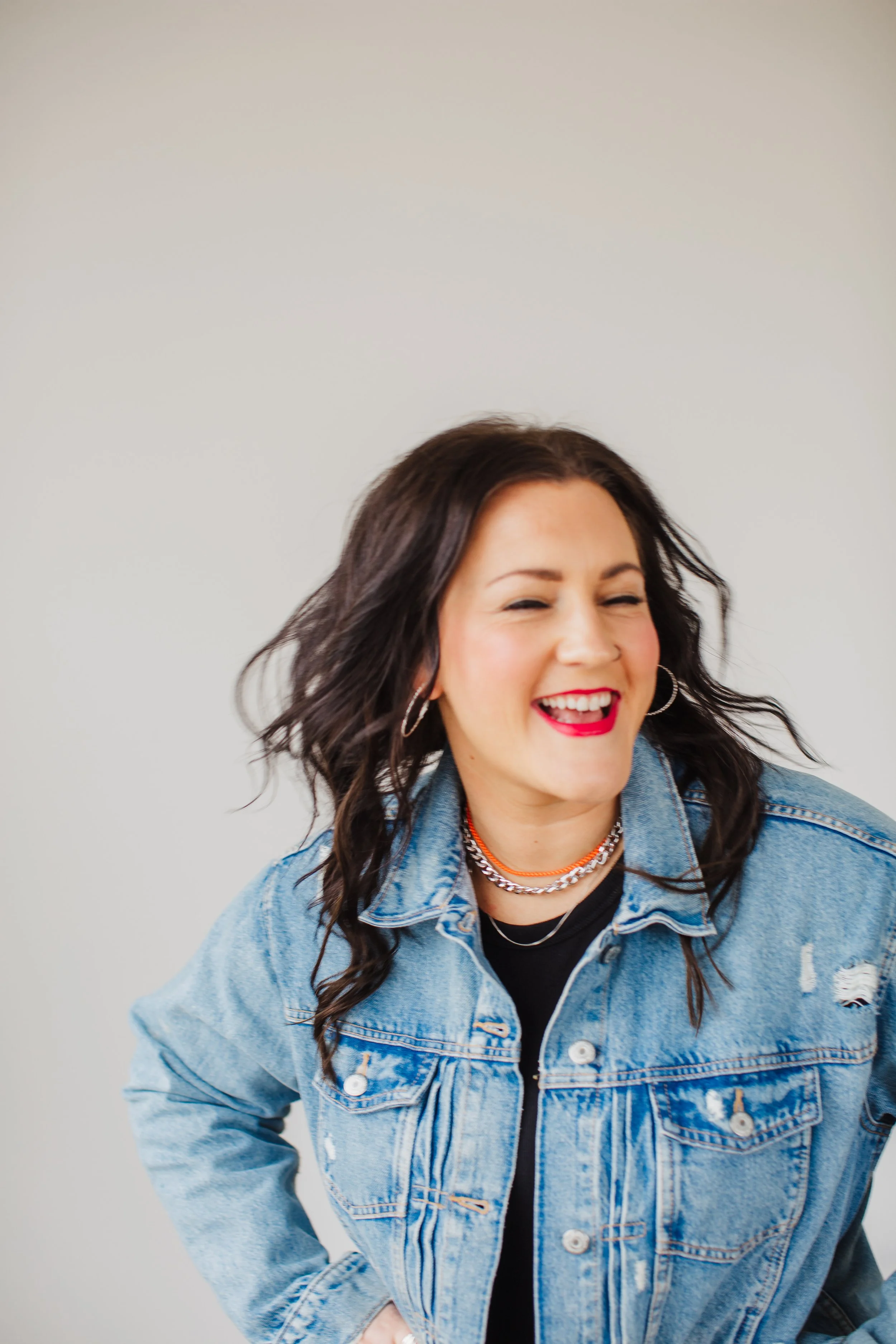 A woman with dark wavy hair smiling, wearing a denim jacket, multiple necklaces, and hoop earrings.