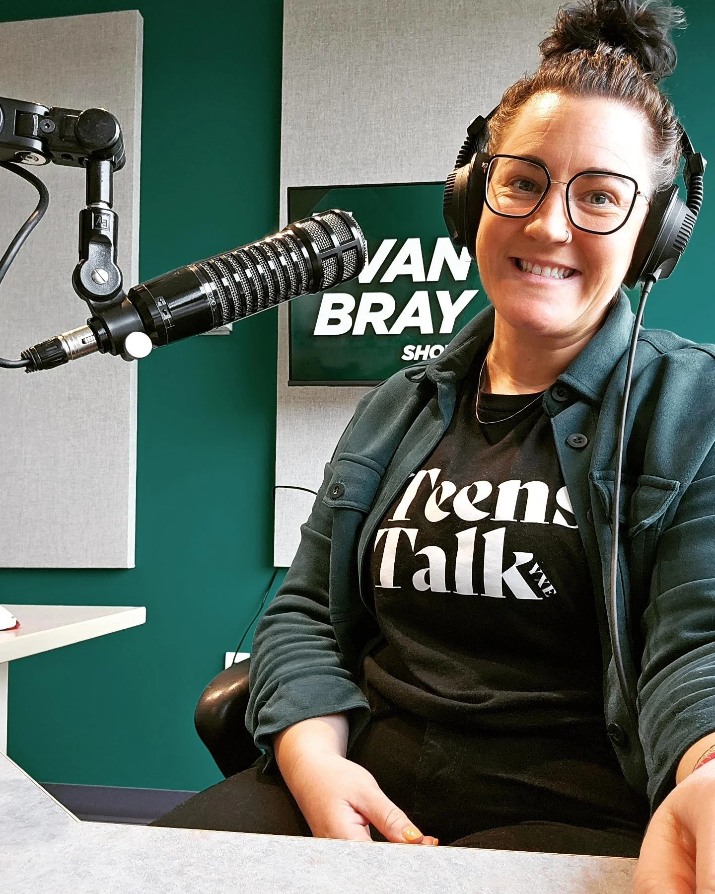 A woman with glasses and a bun hairstyle, smiling, sitting in a radio studio with a microphone and headphones, in front of a screen displaying 'VAN BRAY SHOW'.