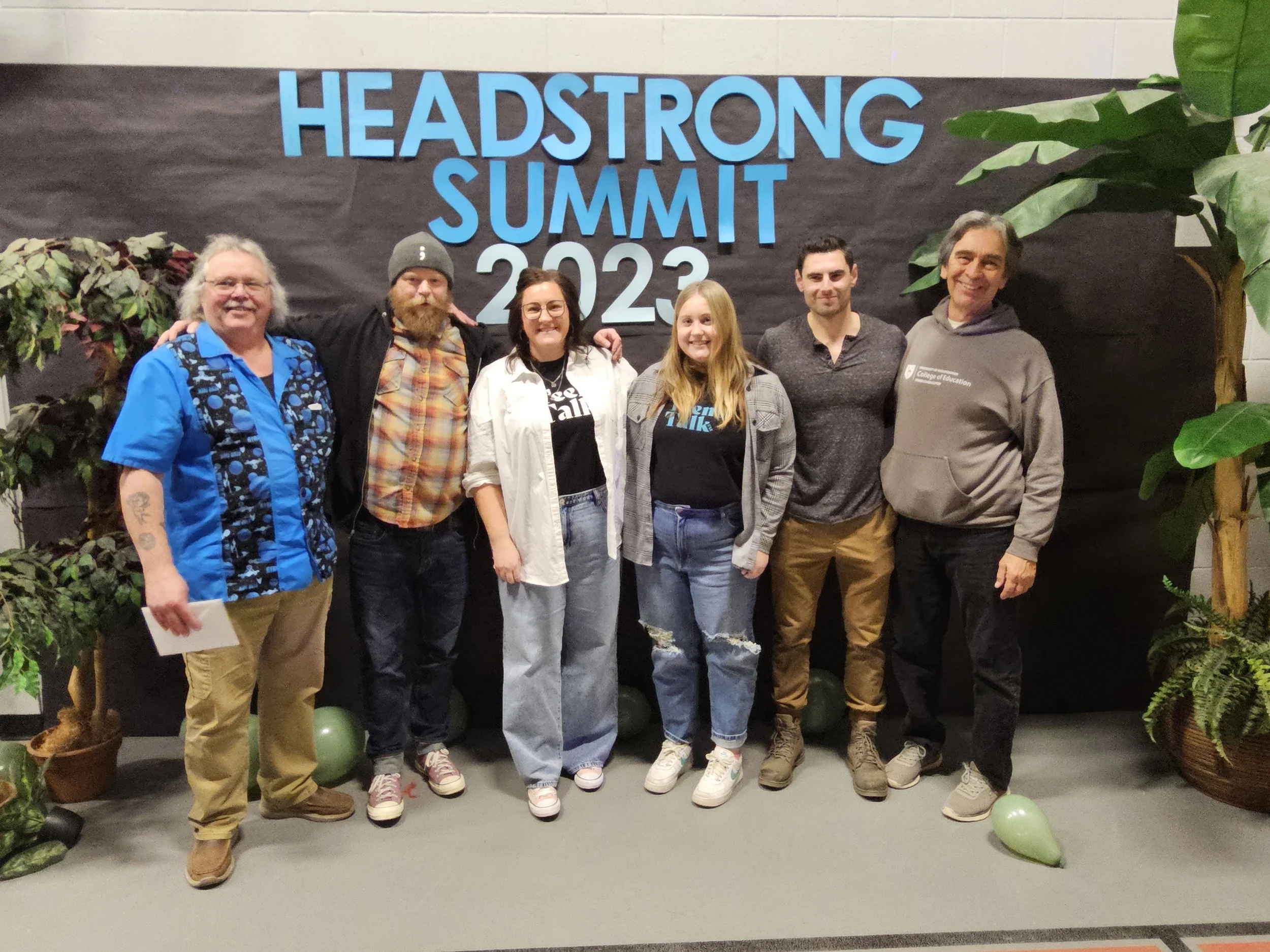 Group of six people standing together and smiling in front of a black backdrop with large blue letters that read 'HEADSTRONG SUMMIT 2023'. Potted plants and green balloons are around them.