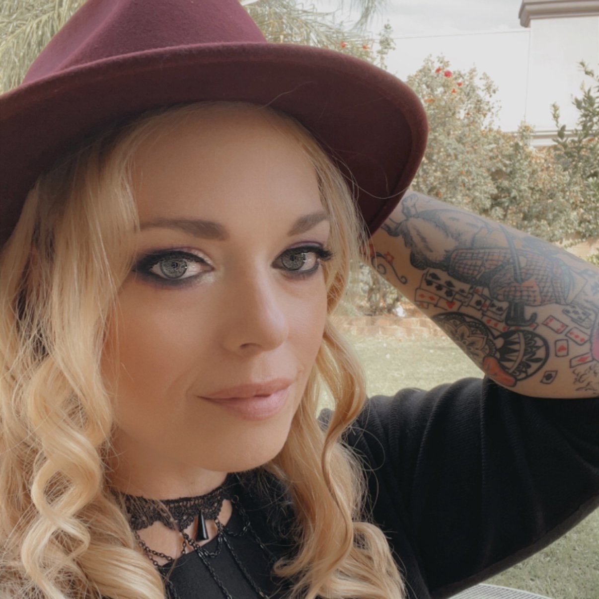 A woman with wavy blonde hair, gray eyes, and a tattooed arm, wearing a maroon hat and a black lace choker, smiling outdoors.