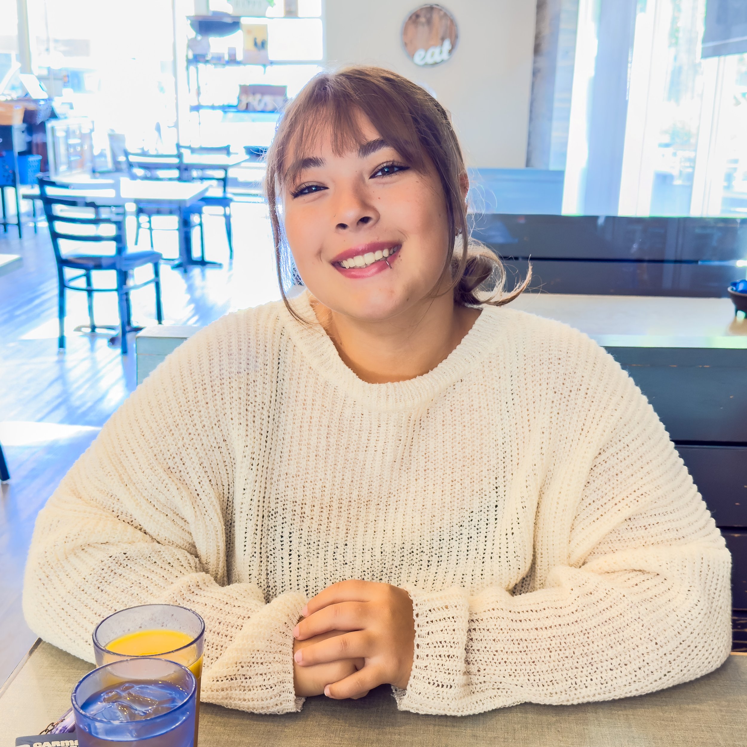 A young woman with light brown hair and bangs, smiling and wearing a cream-colored knitted sweater, sitting at a table in a bright, modern restaurant with large windows and empty tables in the background.