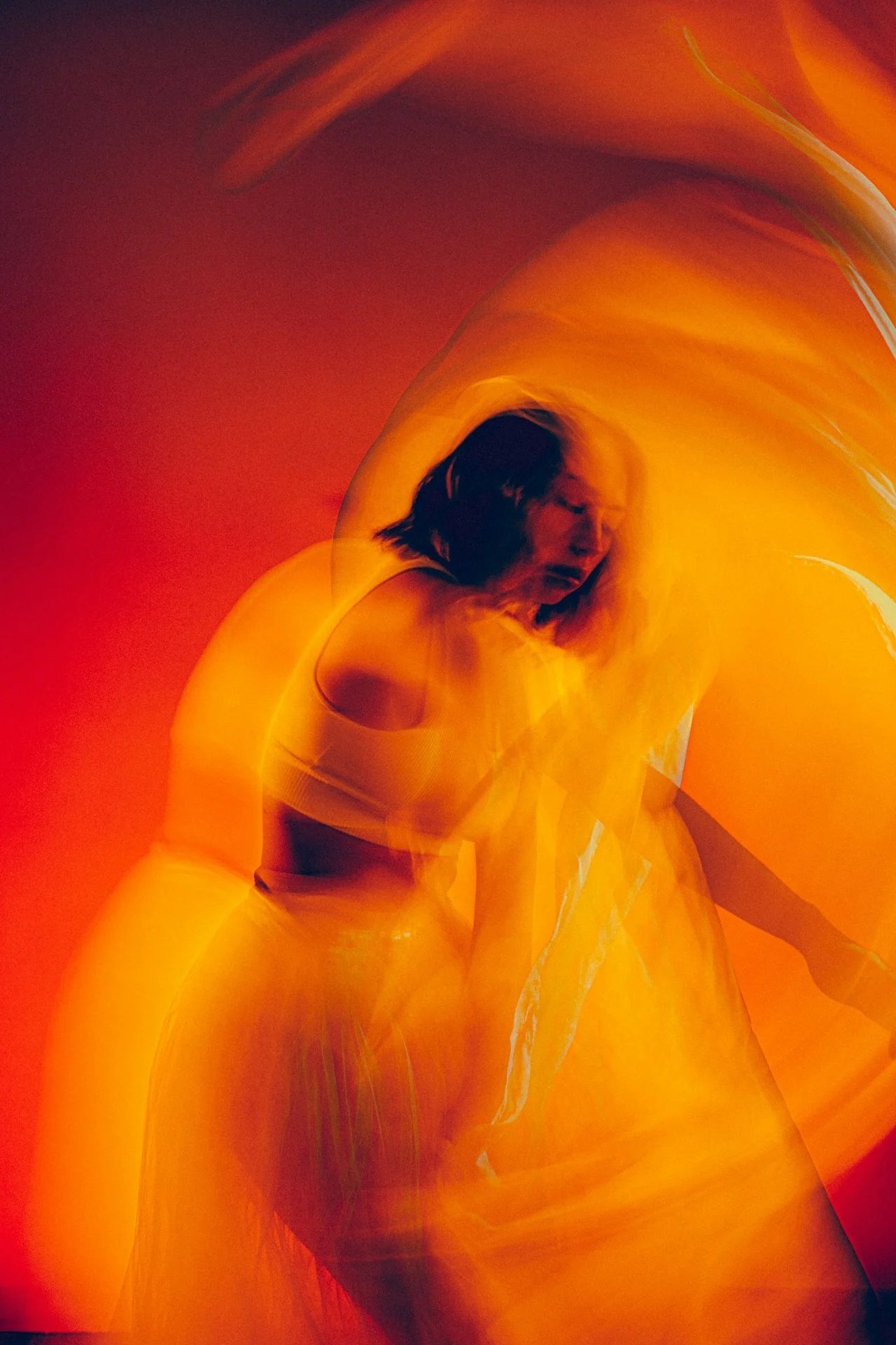 A woman in a white tank top and yellow pants appears to be moving or dancing, with a blurred motion effect in shades of red, orange, and yellow creating an abstract, fiery background.