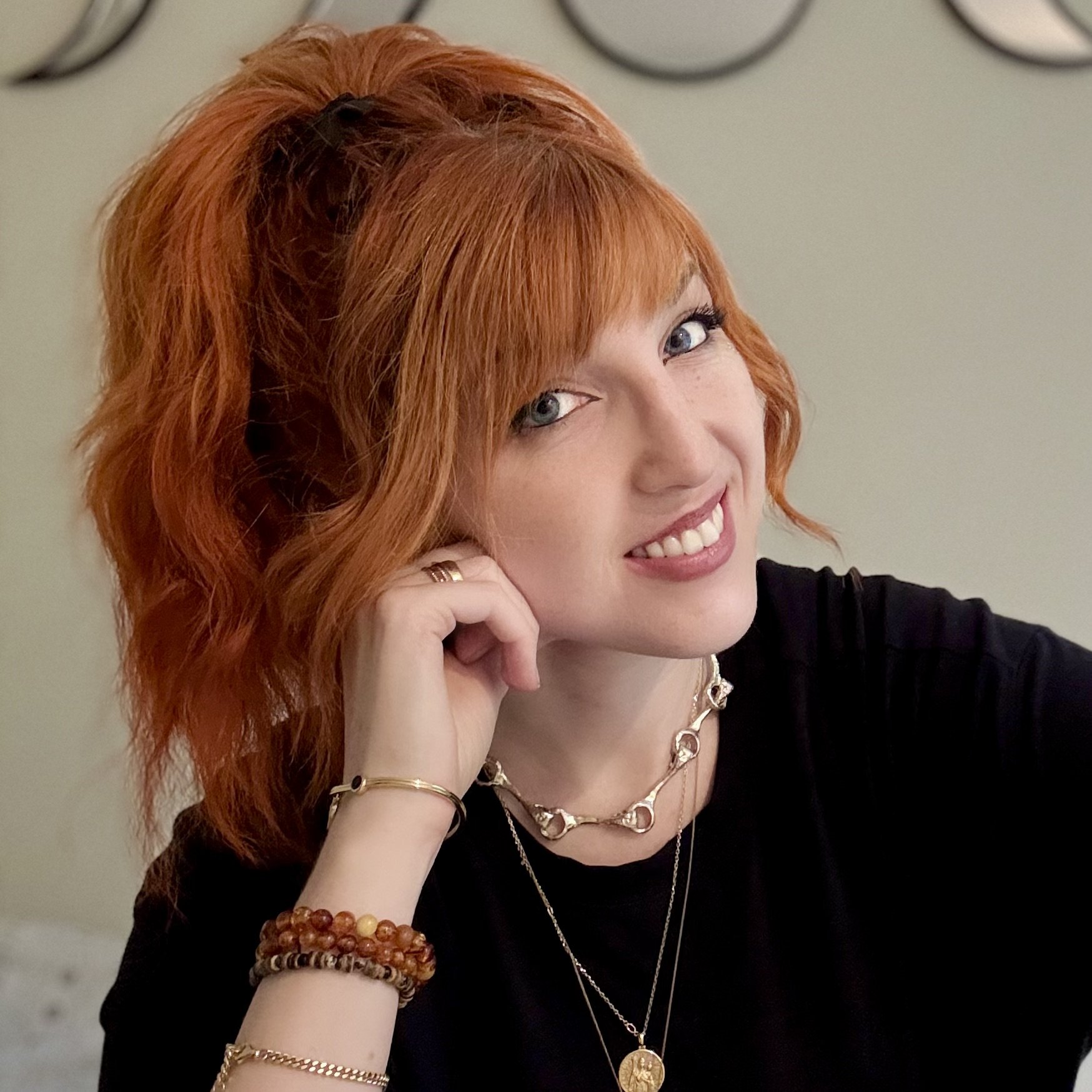 Close-up of a woman with red hair, blue eyes, and a big smile, resting her head on her hand, with jewelry including necklaces, bracelets, and rings, and wearing a black top.