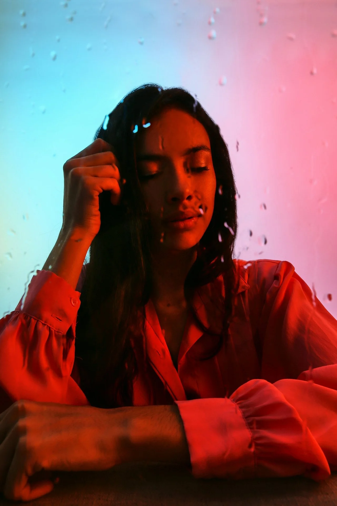 A woman with long dark hair, closed eyes, and a light brown complexion, wearing a satin orange shirt, sitting with her head tilted slightly, in front of a multicolored background with water droplets on a glass or window.