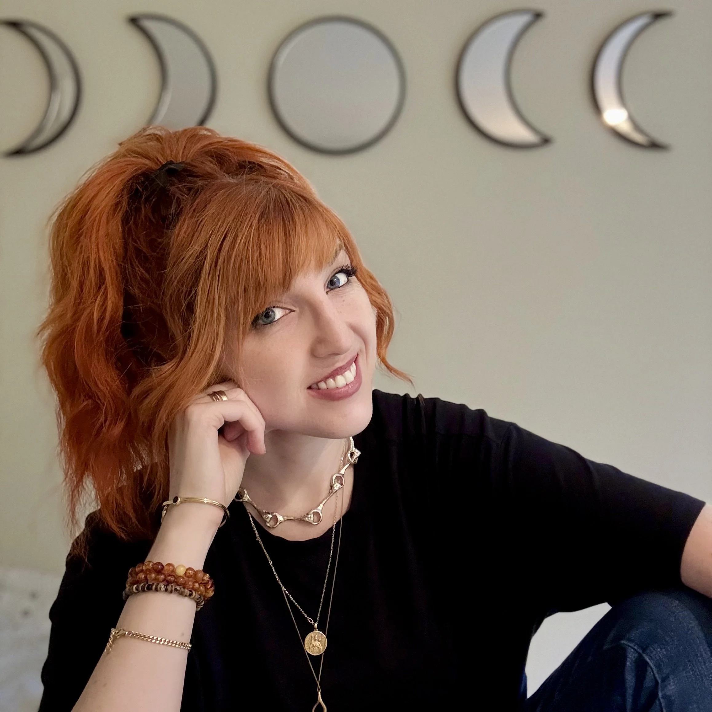 A woman with vibrant red hair smiling and looking at the camera, wearing a black top and layered jewelry, with moon phase wall art in the background.