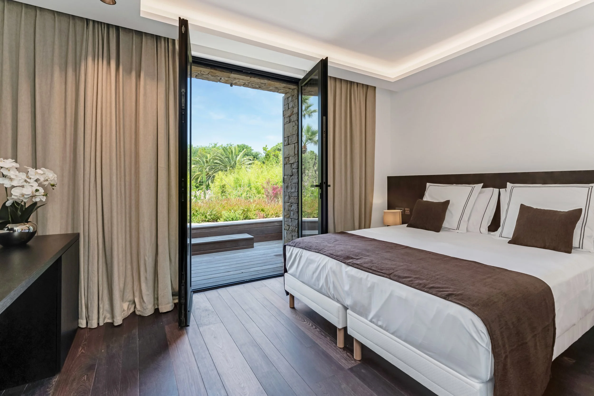 A bedroom with a bed, brown and white bedding, and a small bedside lamp. There are beige curtains and glass doors leading to a small outdoor deck with a view of greenery and trees.