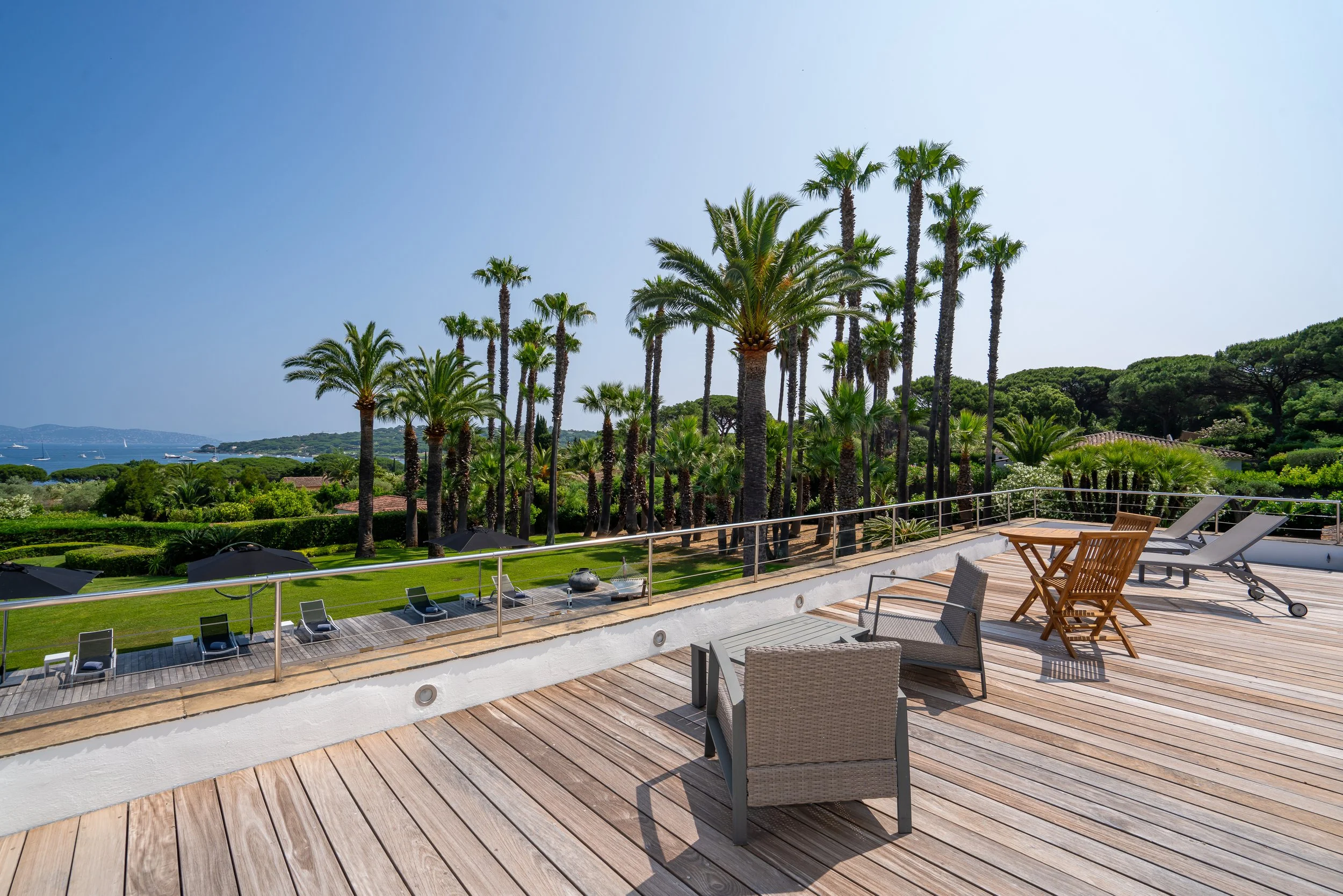 Luxury outdoor patio with wooden deck, lounge chairs, a small table, and a garden with palm trees under a clear blue sky.