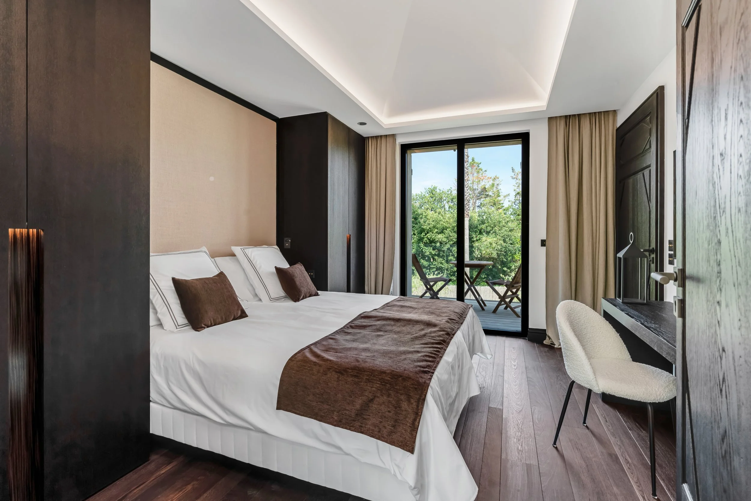 Modern hotel room with a large bed, brown accents, and a balcony with outdoor seating overlooking trees.