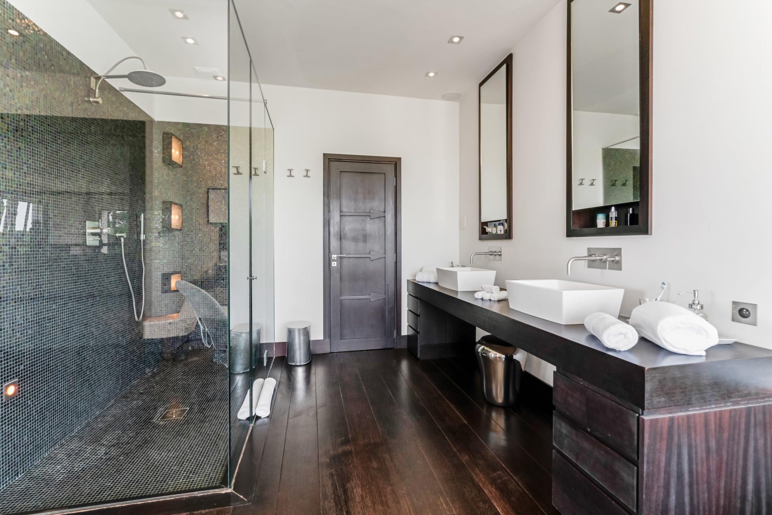 Luxury bathroom with walk-in shower, dark wood floor, double vanity with two white vessel sinks, mirrors, and bathroom essentials.