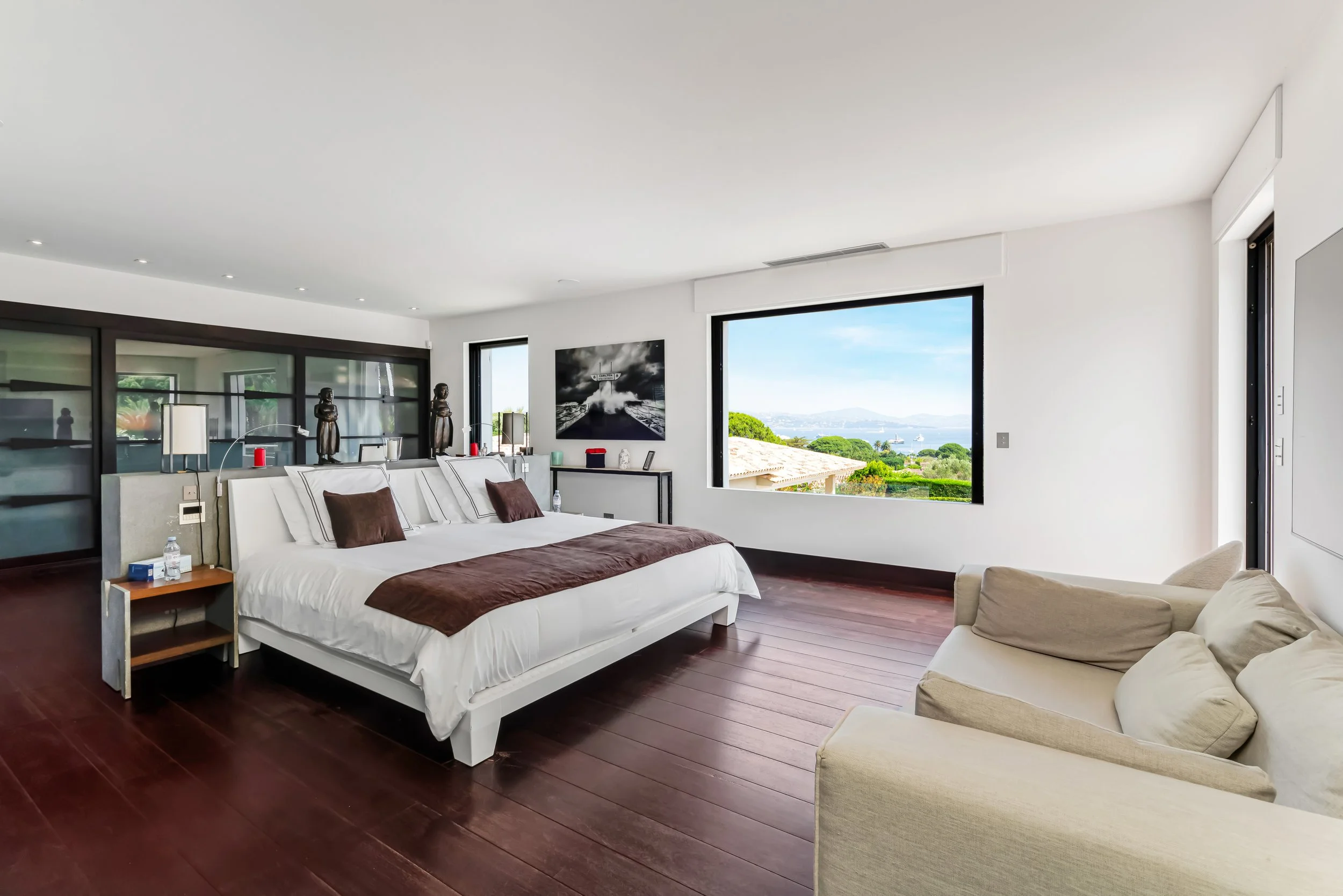 Modern bedroom with large bed, dark wood floors, white walls, large windows overlooking a green landscape and ocean, beige sofa, and black and white artwork.