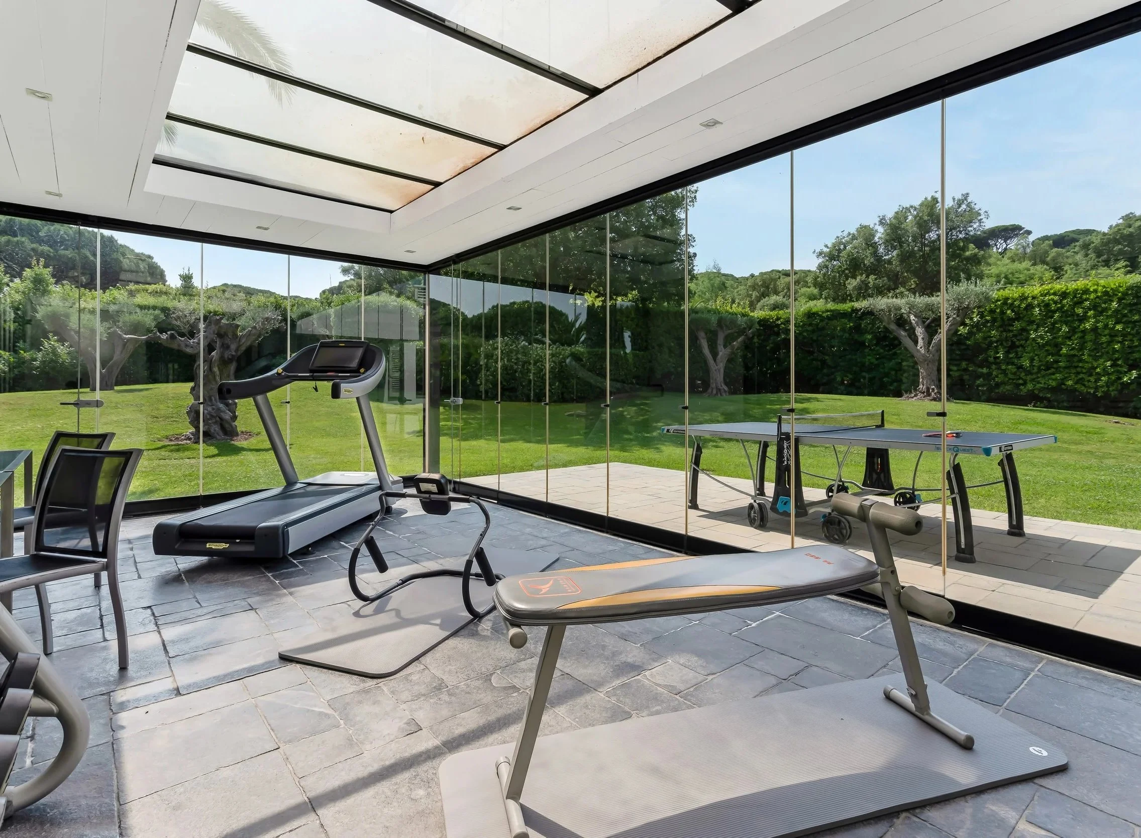 Home gym with treadmill, elliptical, weight bench, and ping pong table, with large glass windows overlooking a green outdoor yard.