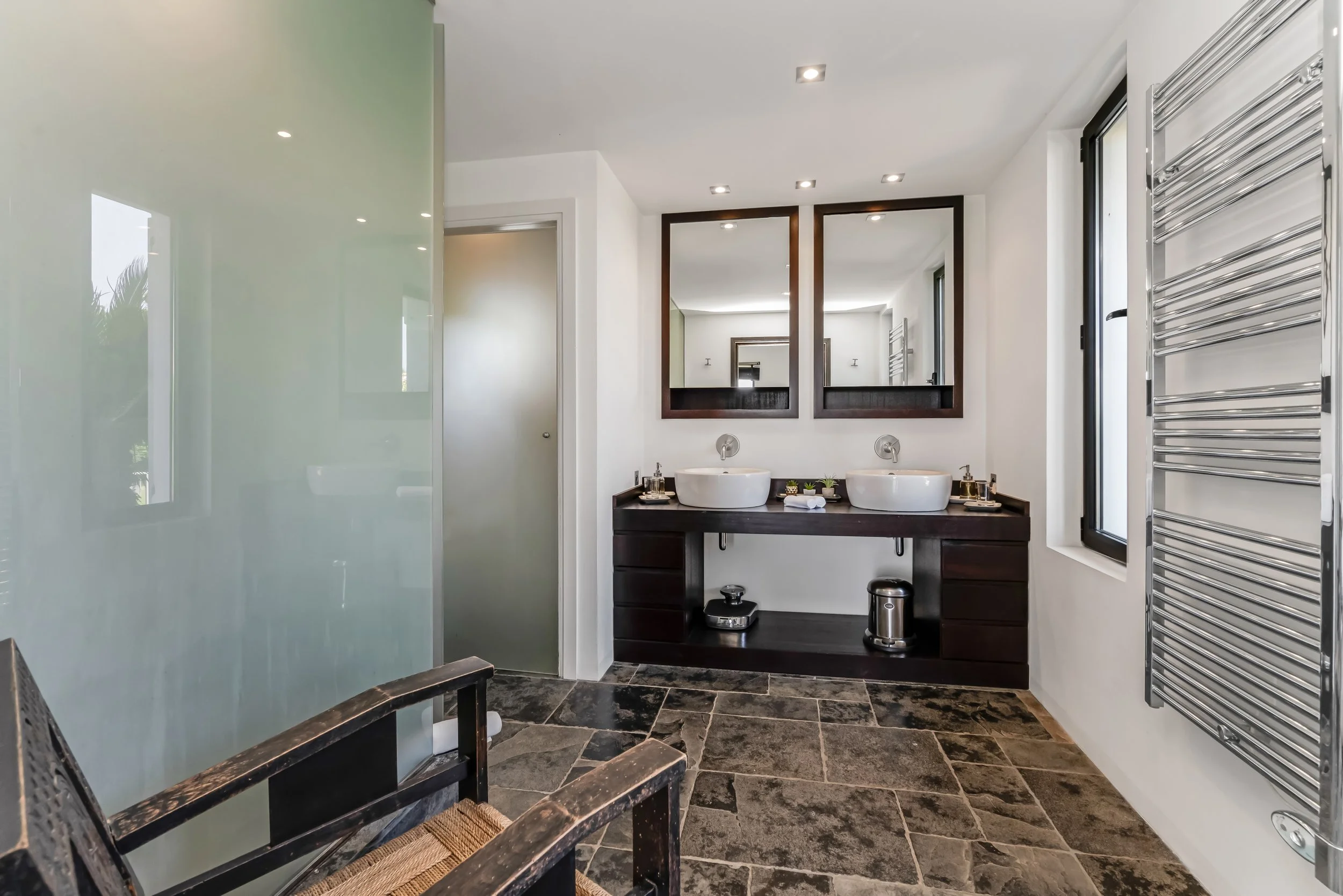 Modern bathroom with double sinks, large mirrors, frosted glass shower, and dark wood vanity, with stone flooring and chrome towel warmer.