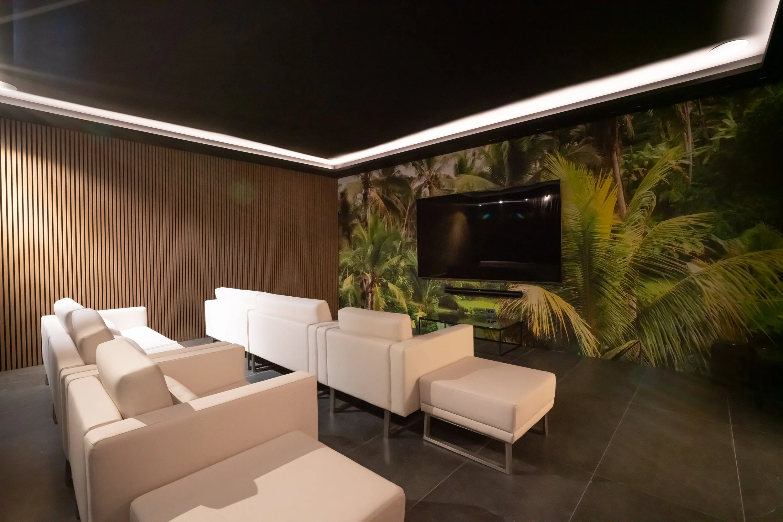Modern living room with white sofas, a large flat-screen TV, tropical wallpaper, and wooden wall paneling.
