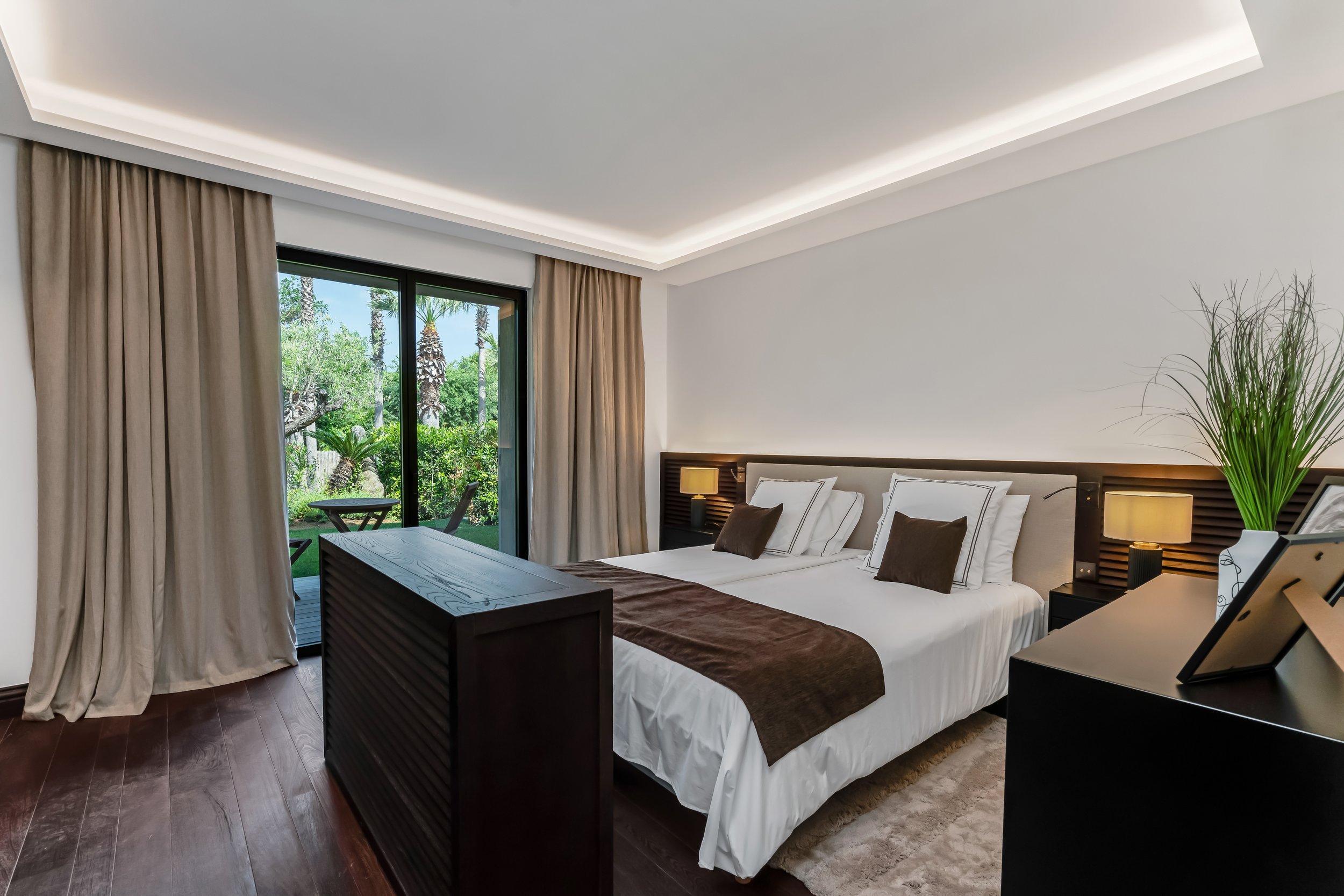 Modern bedroom with a large bed, dark wood furniture, and sliding glass doors leading to a garden patio with trees and greenery.