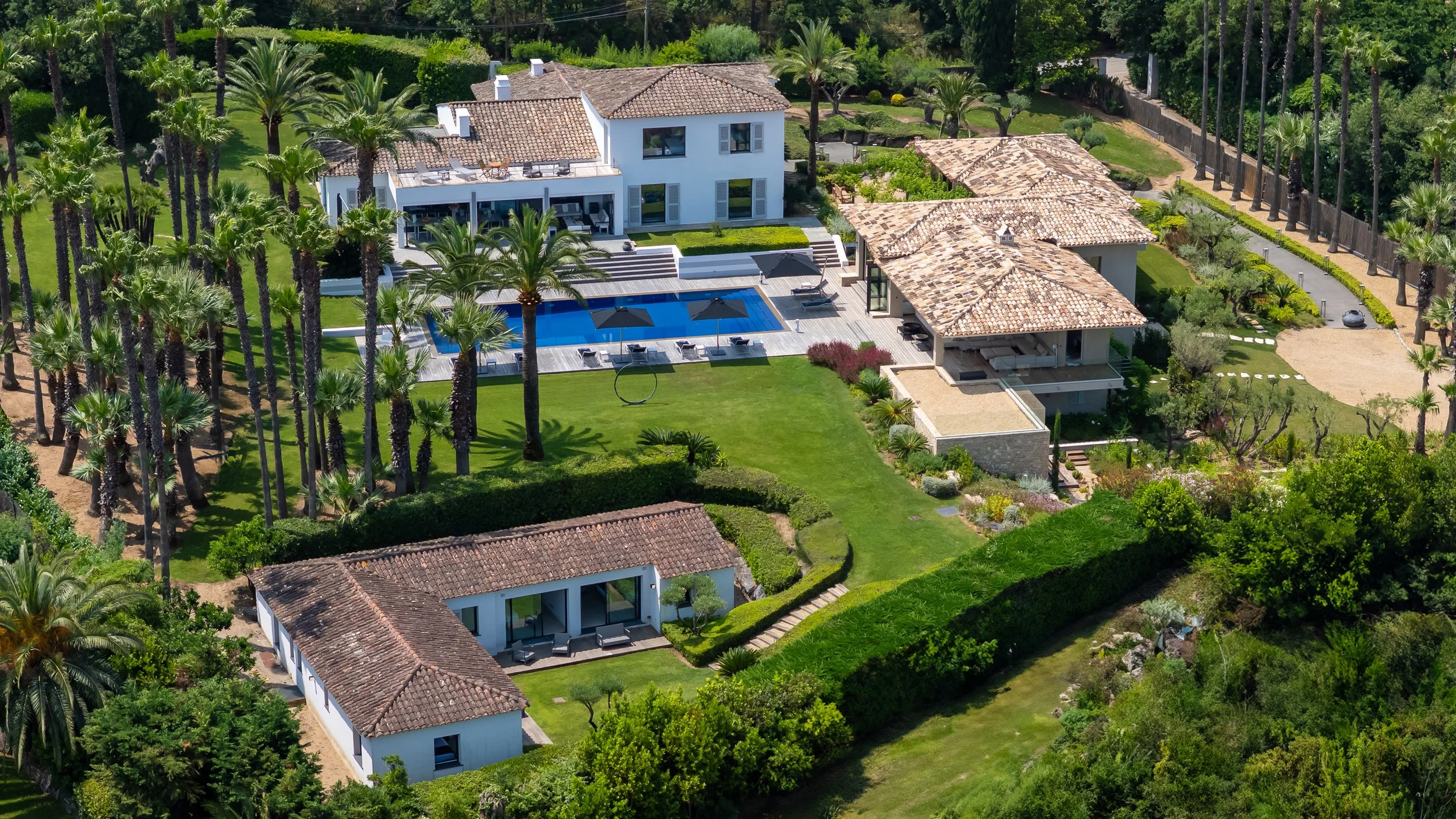 Aerial view of a luxurious estate featuring a white main house with a swimming pool, surrounded by lush landscaped gardens, tall palm trees, and multiple smaller structures with terracotta roofs.