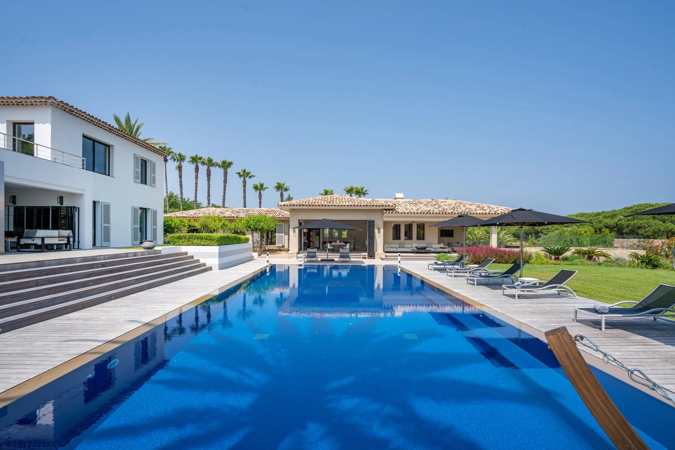 Luxury house with a large swimming pool surrounded by lounge chairs and shaded umbrellas, with a lush green landscape and palm trees in the background under a clear blue sky.