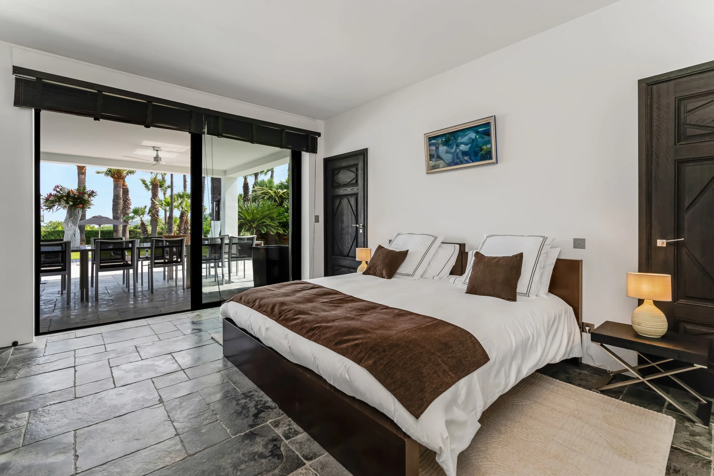 Modern bedroom with large bed and white linens, brown blanket, and pillows, with sliding door leading to outdoor patio with dining table and chairs, surrounded by palm trees.